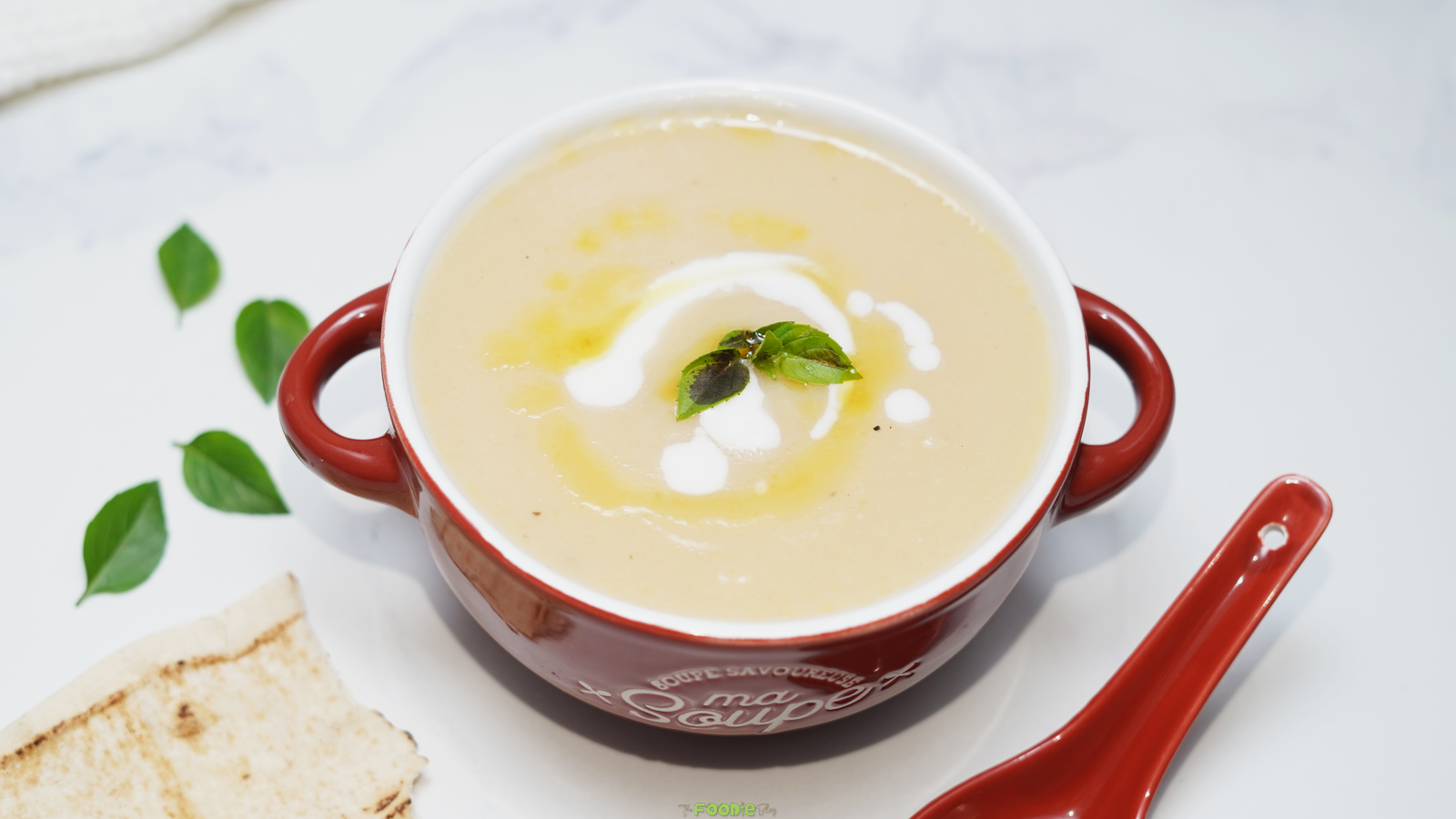 Overhead view of creamy cauliflower soup in a red handled bowl with cream, olive oil, and basil garnish.