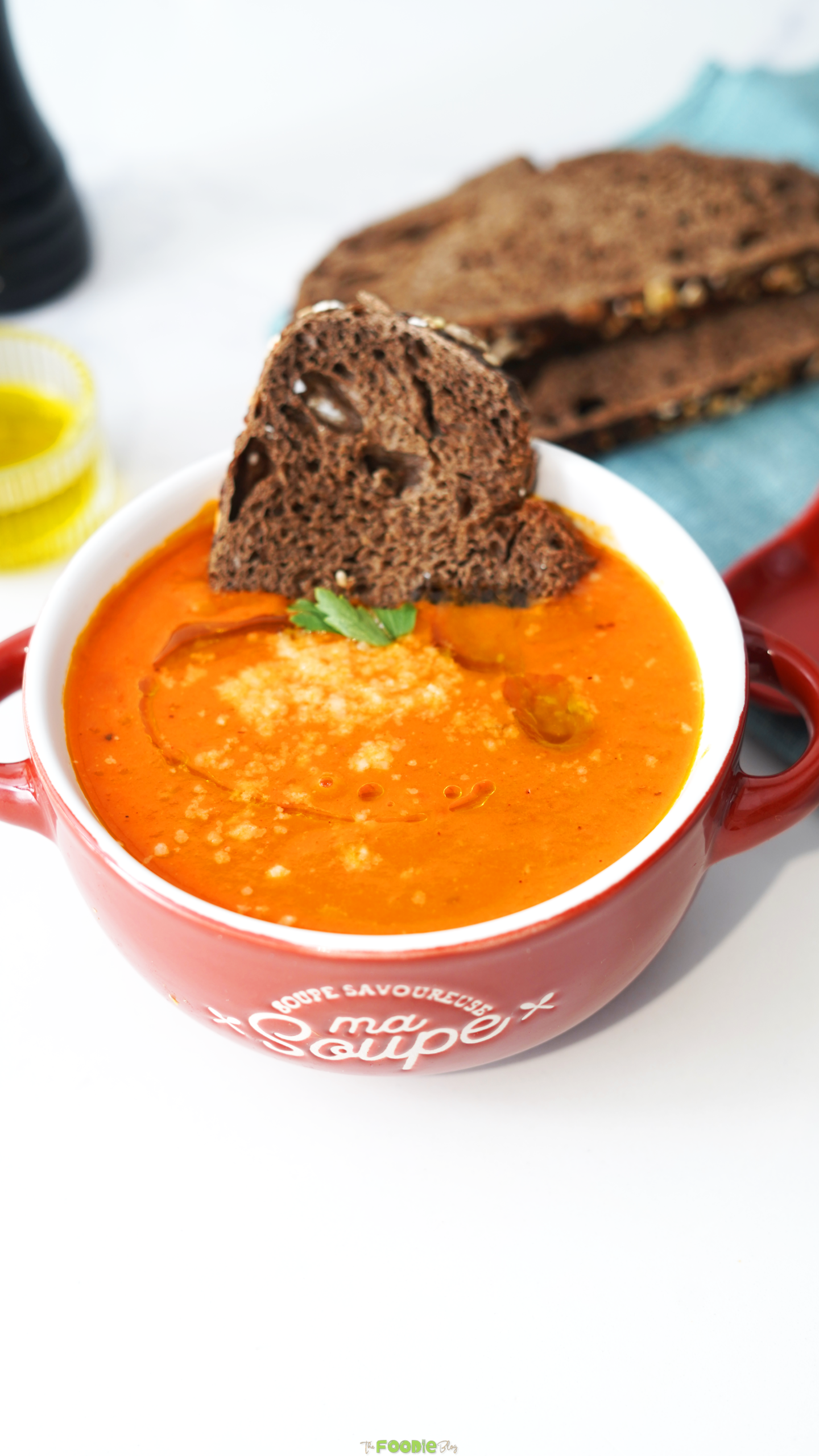 Red bowl of creamy tomato soup topped with parmesan, olive oil, parsley, and toasted bread.