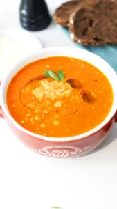 Bowl of creamy tomato soup topped with parmesan, olive oil, and a fresh herb leaf.