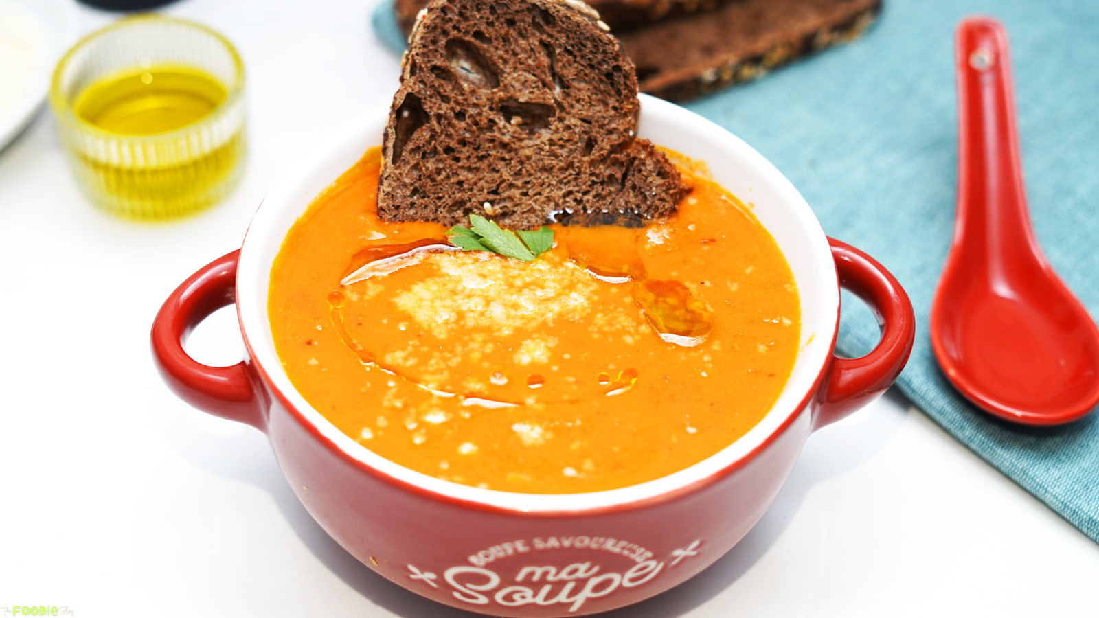 Creamy tomato soup in a red bowl topped with parmesan, olive oil, and a slice of toasted bread.