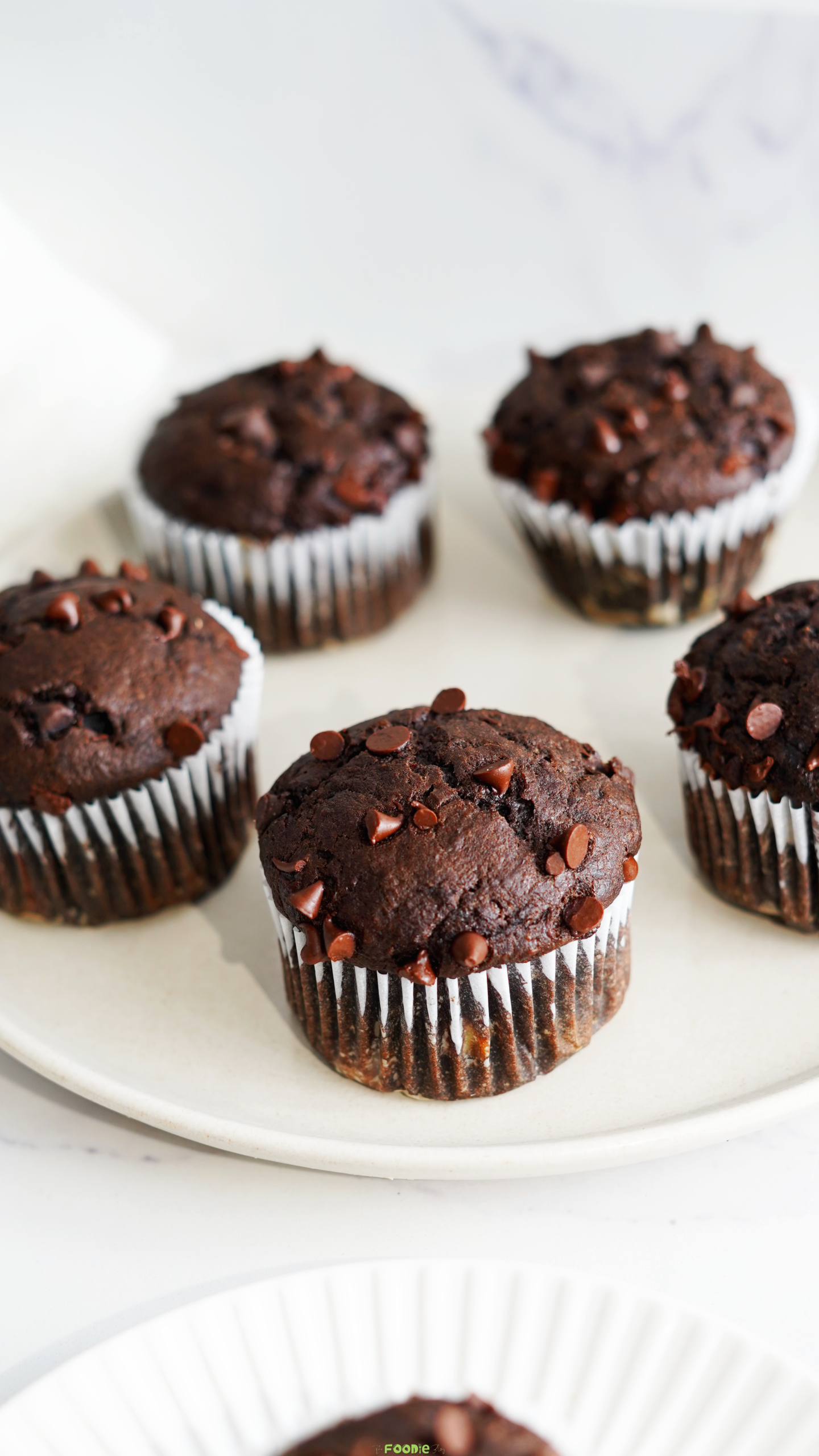 Freshly baked double chocolate protein muffins with chocolate chips on top arranged on a white plate.
