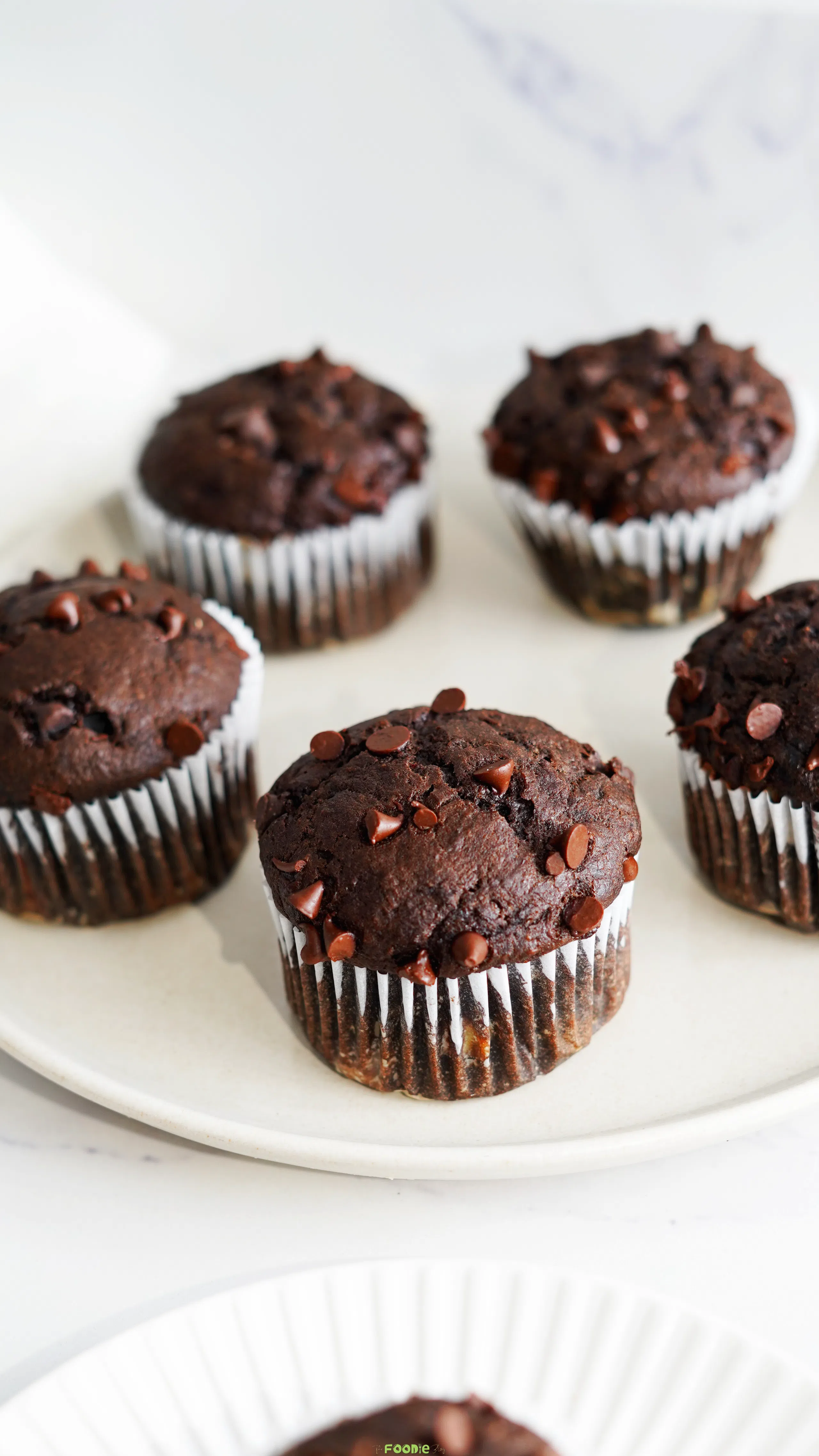 Freshly baked double chocolate protein muffins with chocolate chips on top arranged on a white plate.
