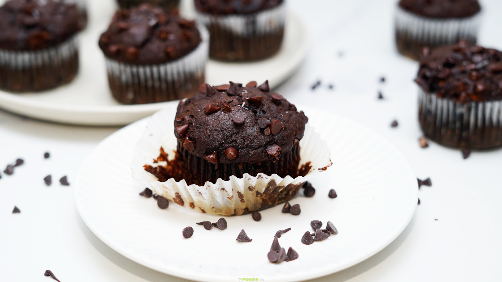 Double chocolate protein muffin on a white plate with more muffins in the background and chocolate chips scattered around.