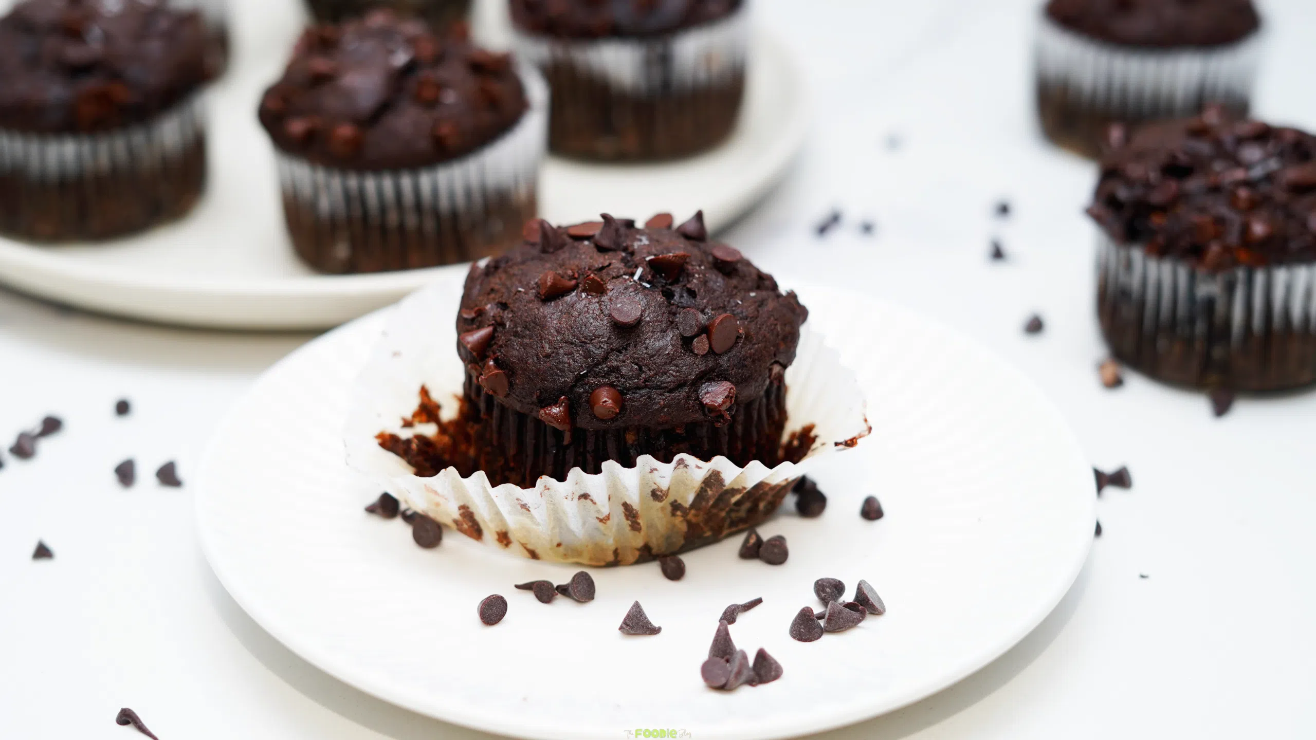 Double chocolate protein muffin on a white plate with more muffins in the background and chocolate chips scattered around.