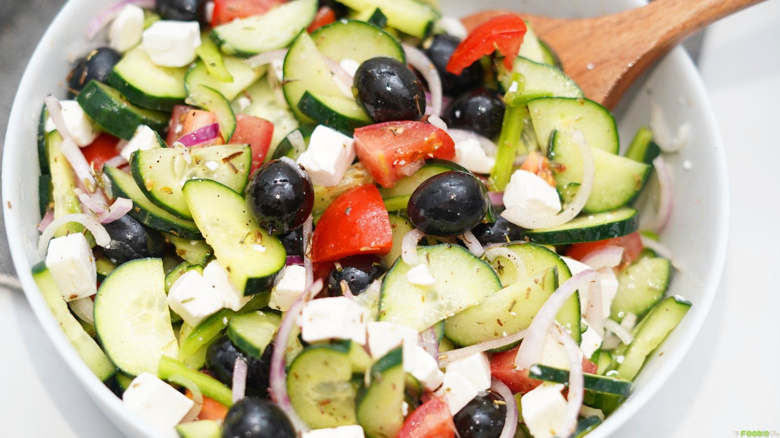 Close-up of Greek Salad with sliced cucumber, tomato chunks, feta cubes, Kalamata olives, red onion, green bell pepper, and oregano dressing.