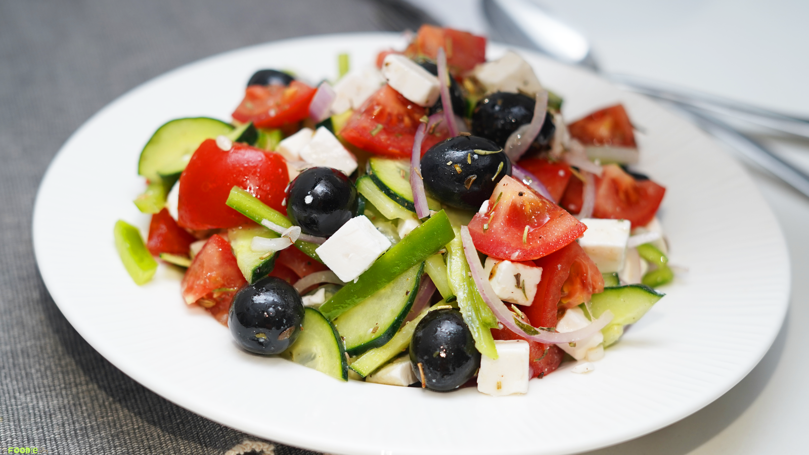 Greek Salad served on a white plate with tomatoes, cucumbers, green bell pepper, feta cubes, Kalamata olives, red onion, and oregano.