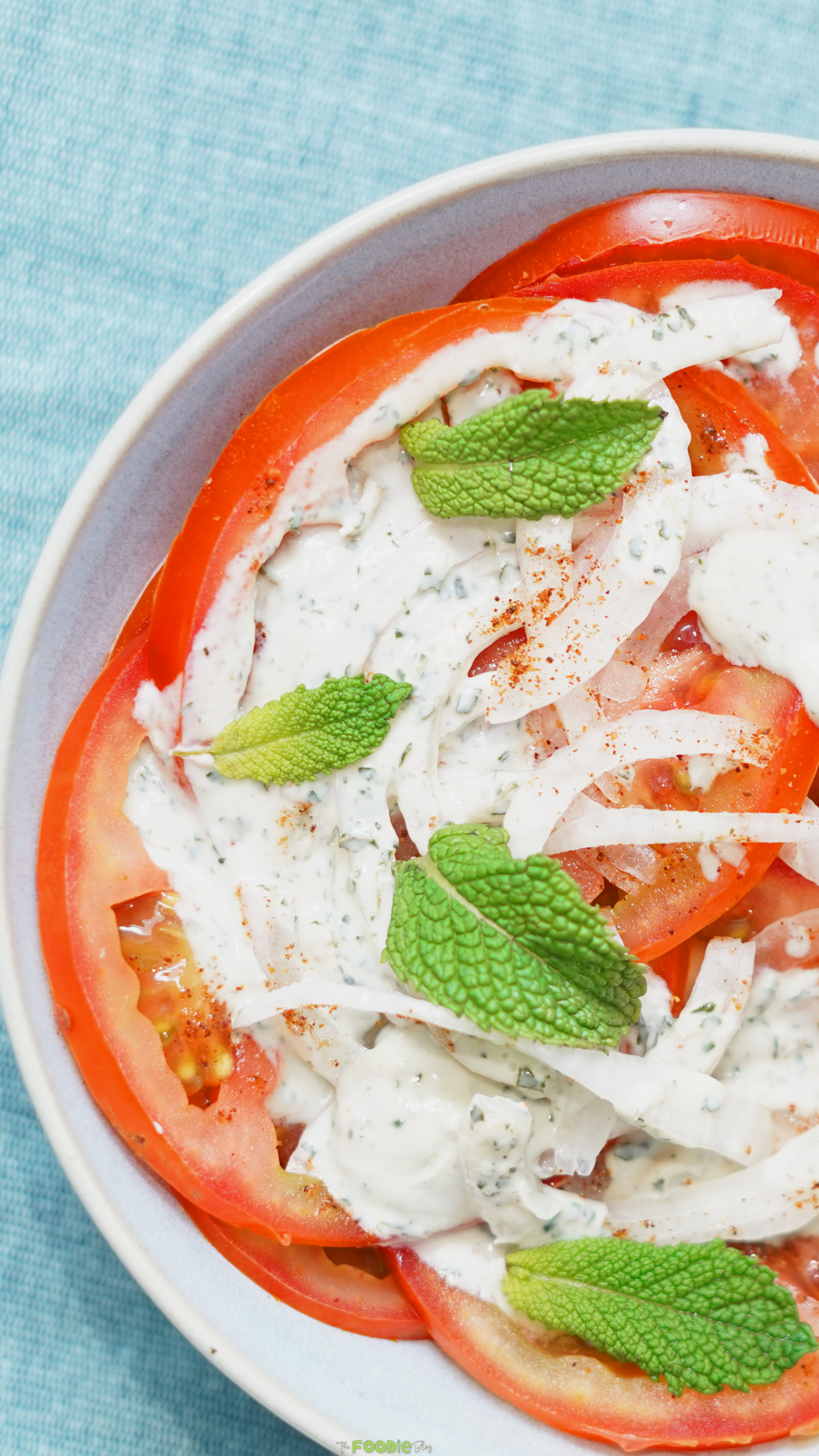 Close-up of sliced tomatoes topped with creamy mint yogurt dressing, thin red onion, fresh mint leaves, and a pinch of spice
