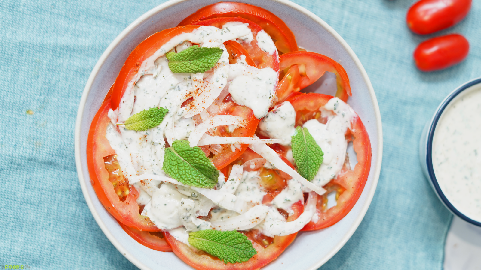 Horizontal photo of sliced tomato salad with creamy mint dressing, red onion, mint leaves, and grape tomatoes nearby.