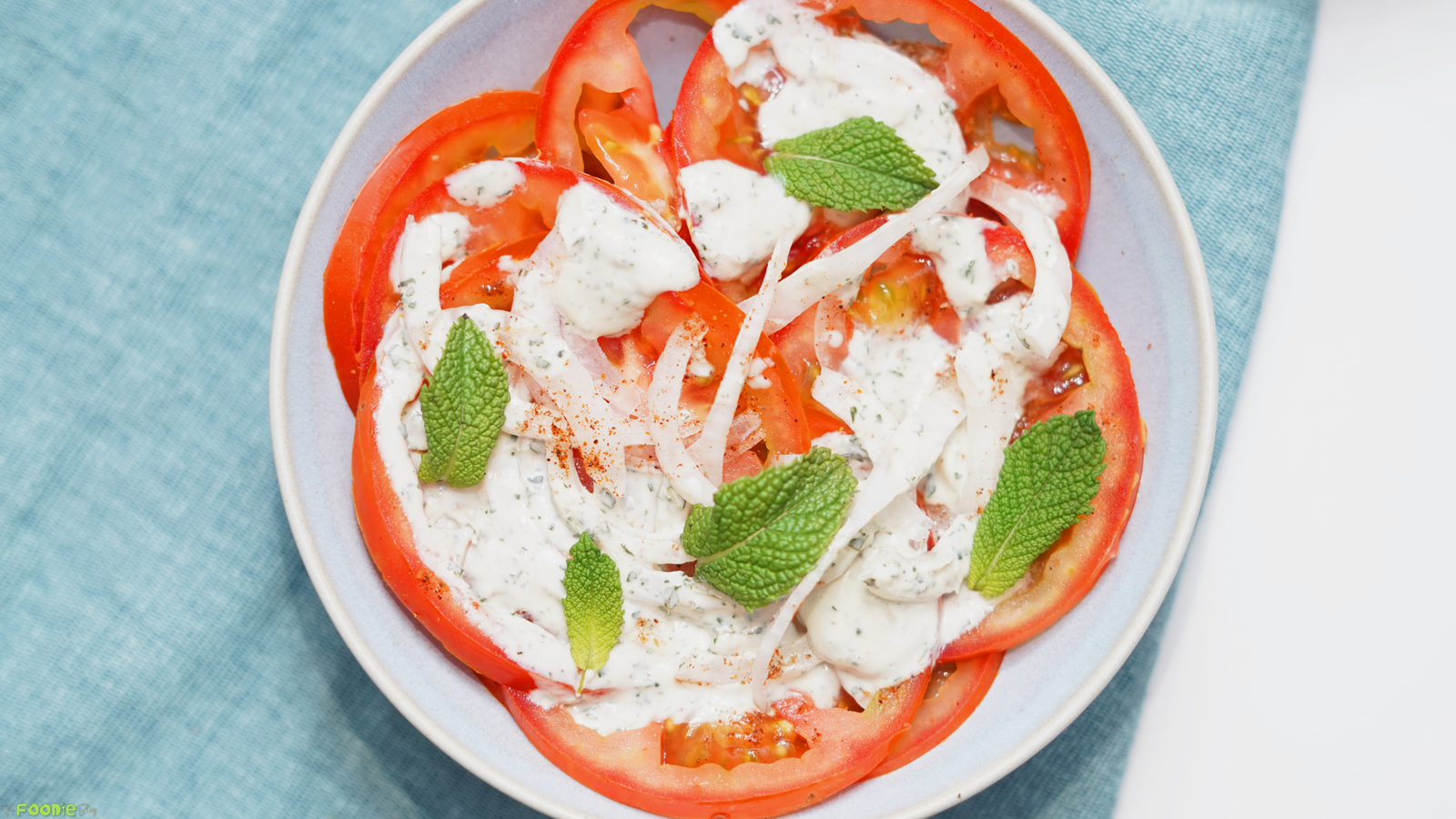 Overhead view of tomato slices on a plate with mint yogurt dressing, red onion, fresh mint, and a light sprinkle of spice.