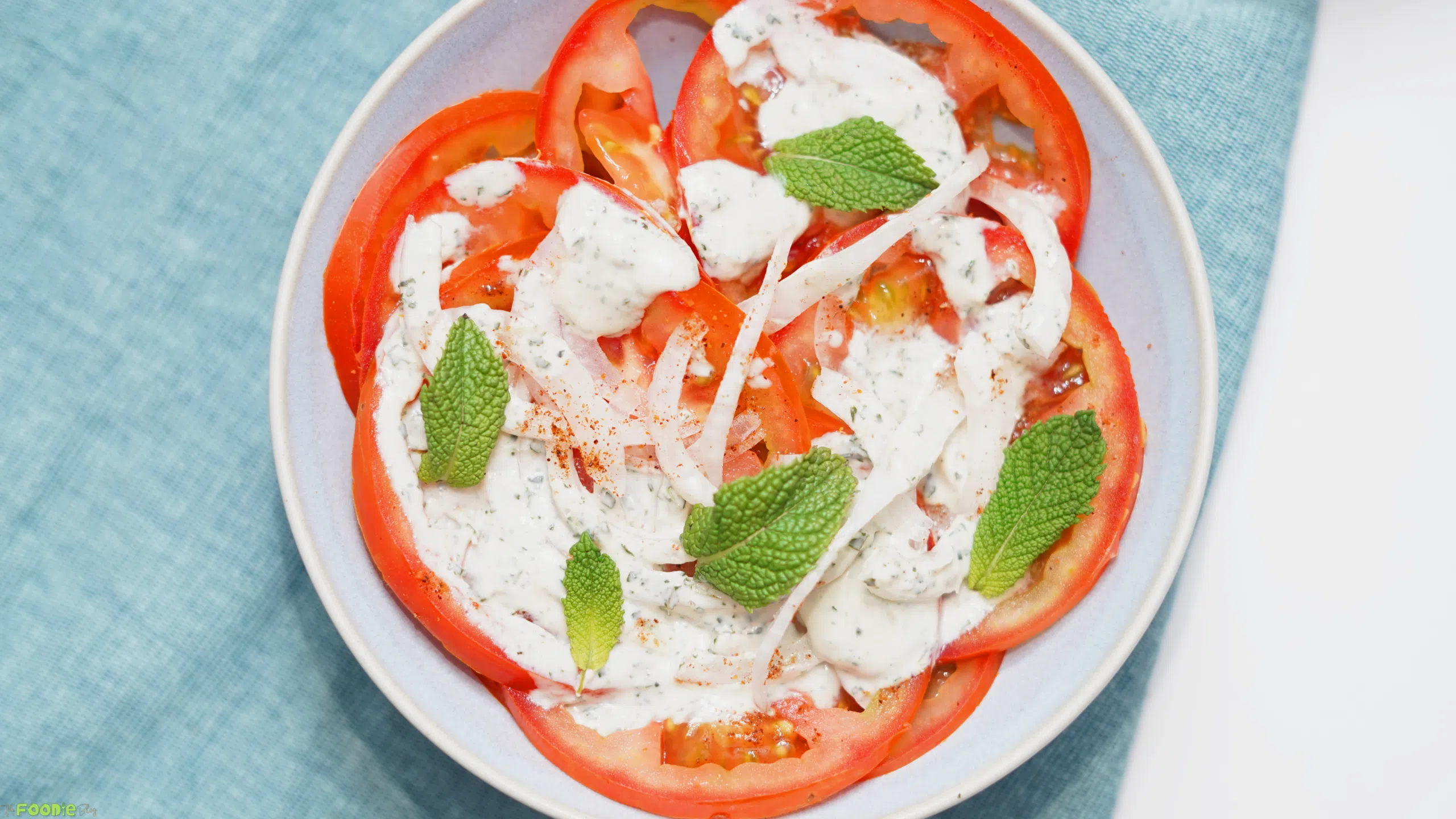 Overhead view of tomato slices on a plate with mint yogurt dressing, red onion, fresh mint, and a light sprinkle of spice.