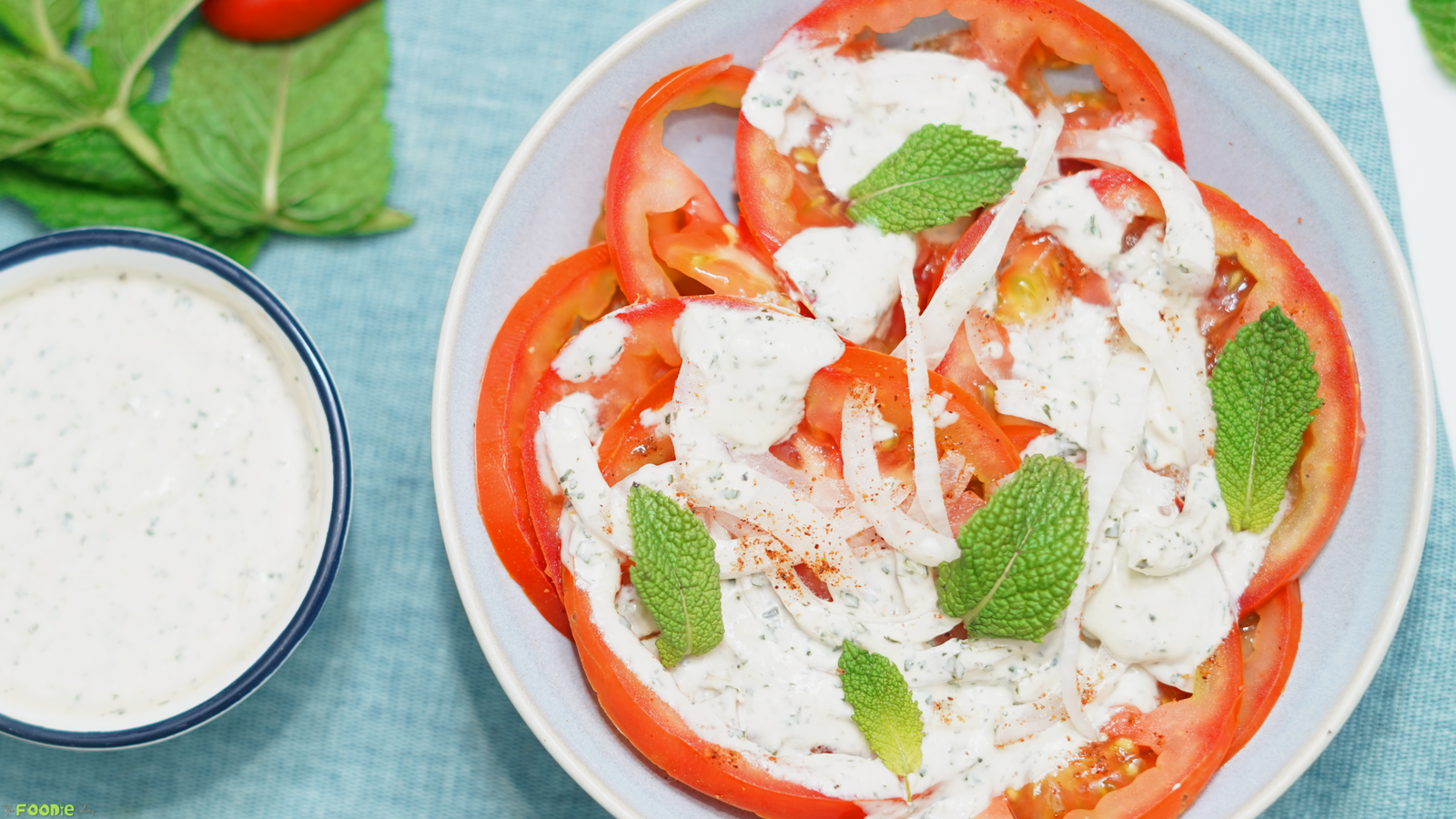 A plate of Indian tomato salad with mint yogurt dressing beside a small bowl of extra dressing on a blue cloth.