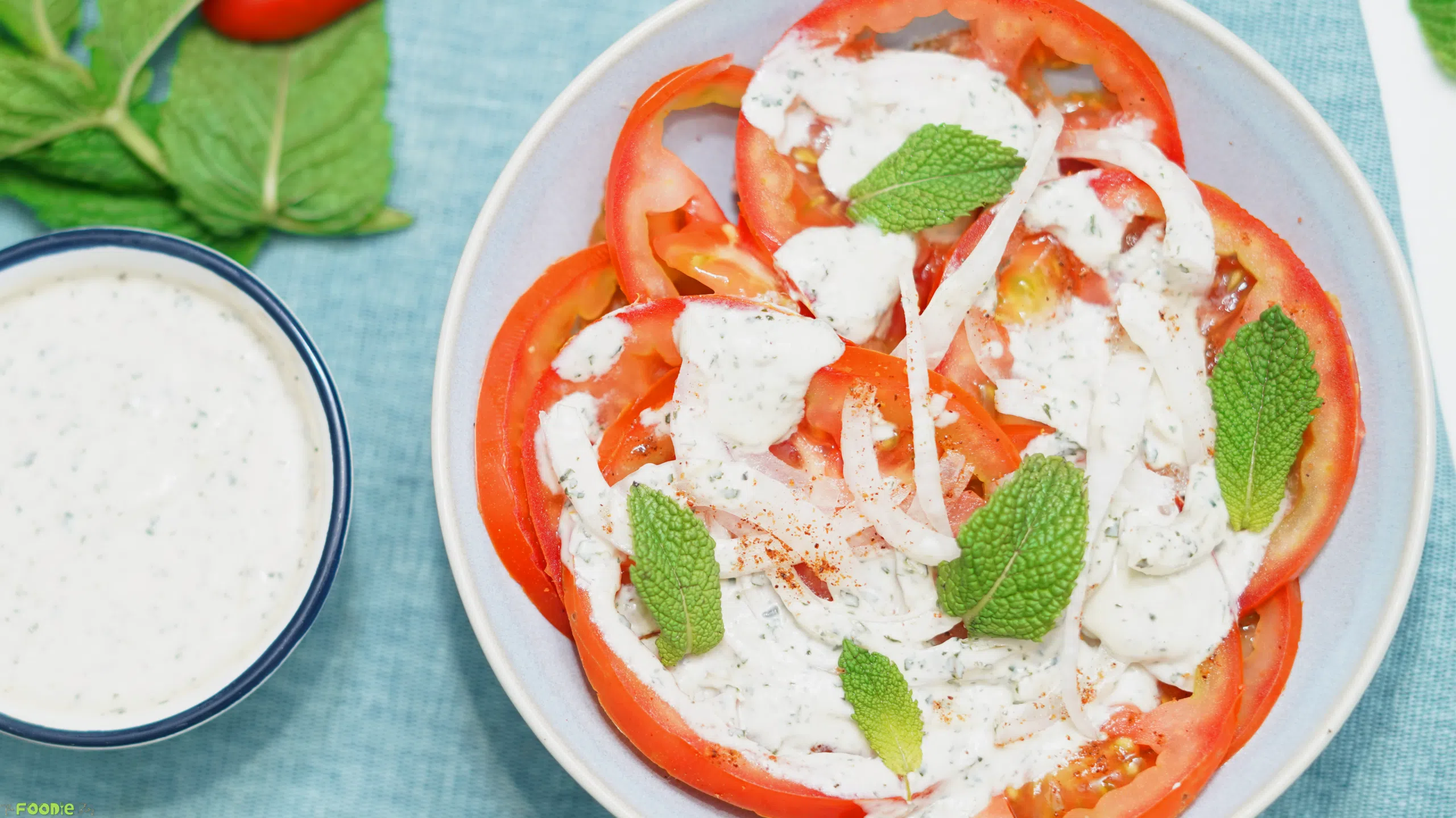 A plate of Indian tomato salad with mint yogurt dressing beside a small bowl of extra dressing on a blue cloth.