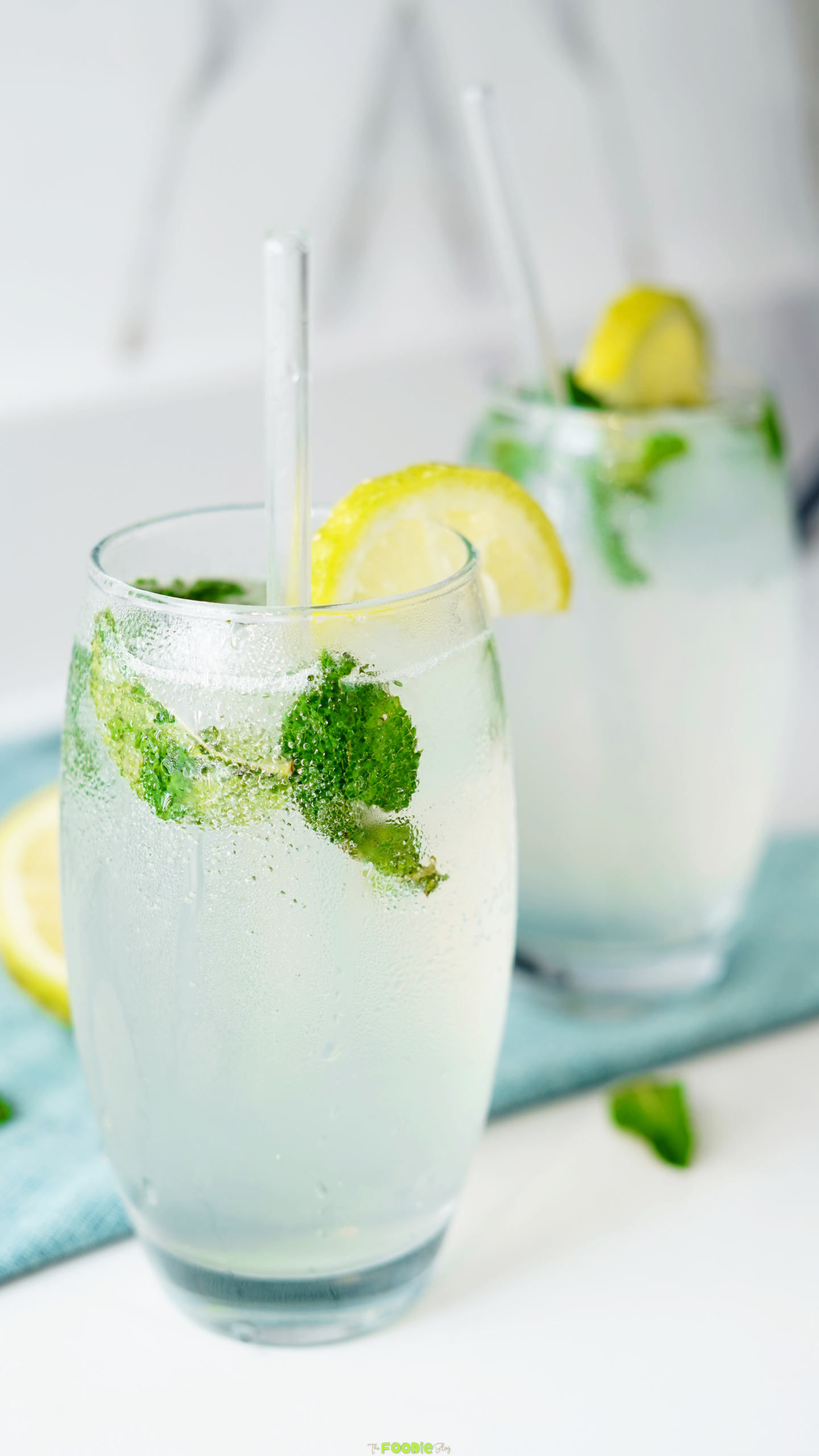 Two mojito drinks with mint leaves, ice, lemon slices, and glass straws on a blue cloth.
