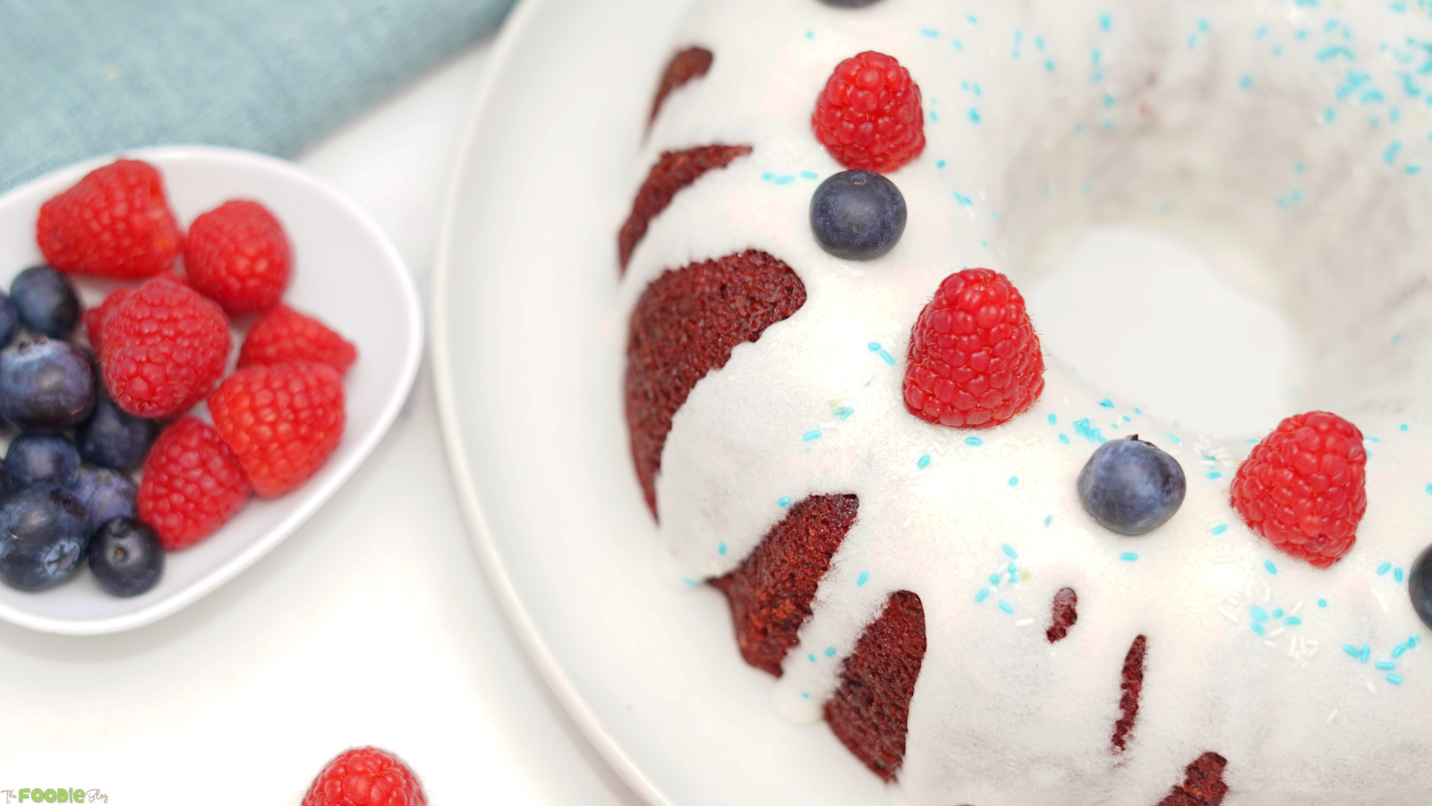 Red velvet Bundt cake with white frosting, raspberries, blueberries, and sprinkles.