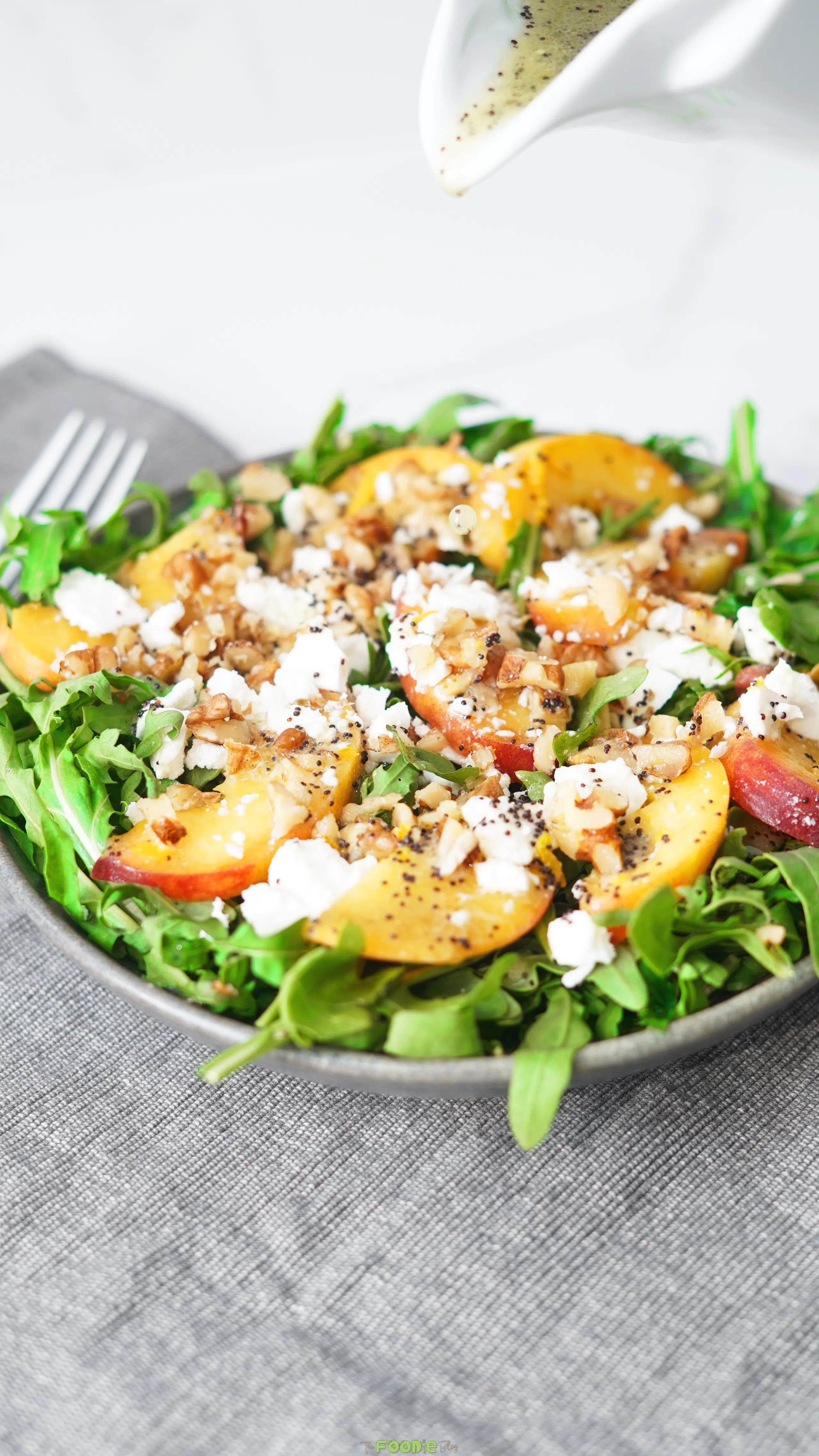 Poppyseed dressing being drizzled over peach salad with arugula, feta, and walnuts.