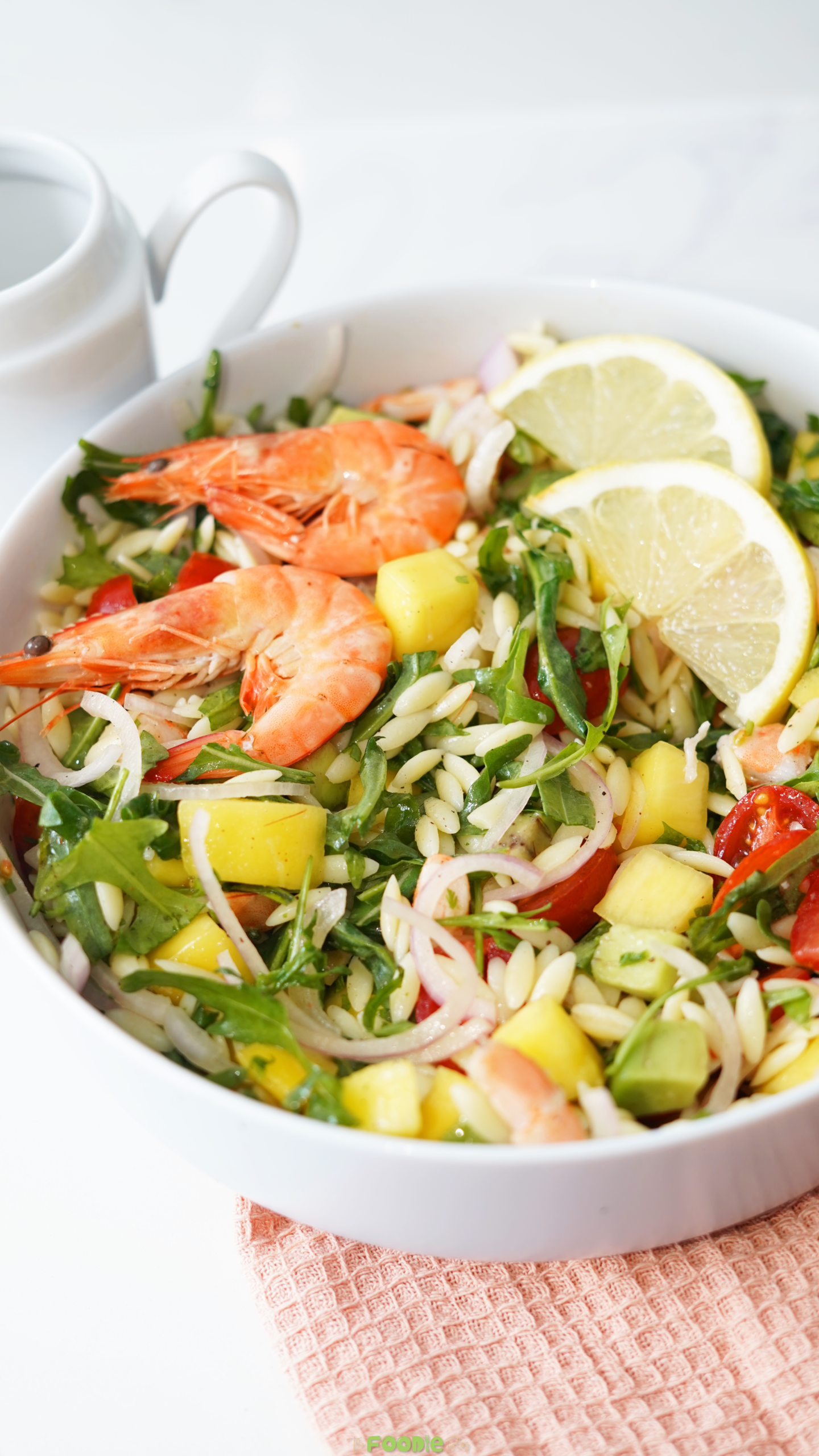 Prawn mango avocado summer salad in a white bowl with lemon slices, shrimp, mango, avocado, orzo, arugula, tomatoes, and red onion.