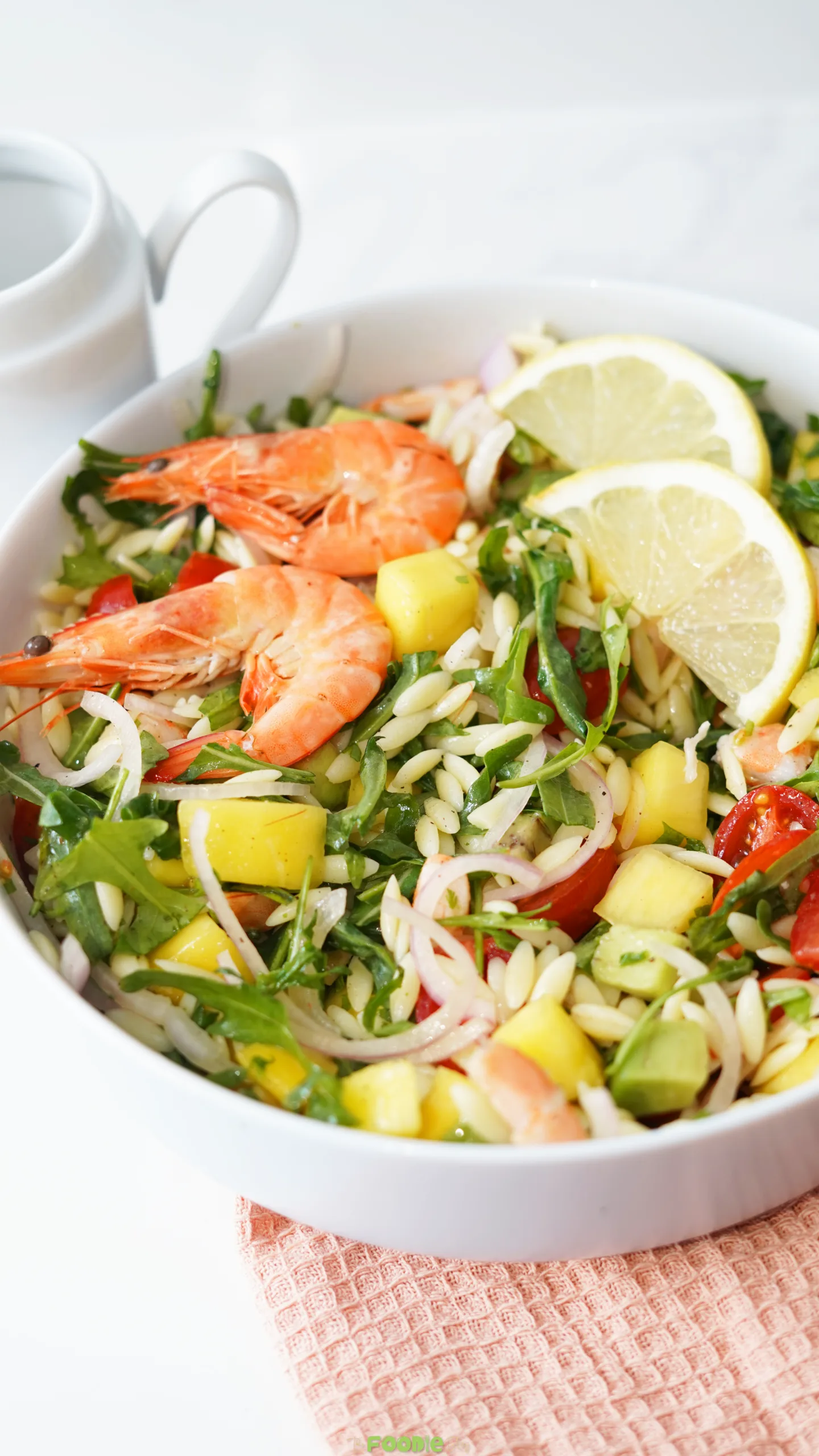 Prawn mango avocado summer salad in a white bowl with lemon slices, shrimp, mango, avocado, orzo, arugula, tomatoes, and red onion.