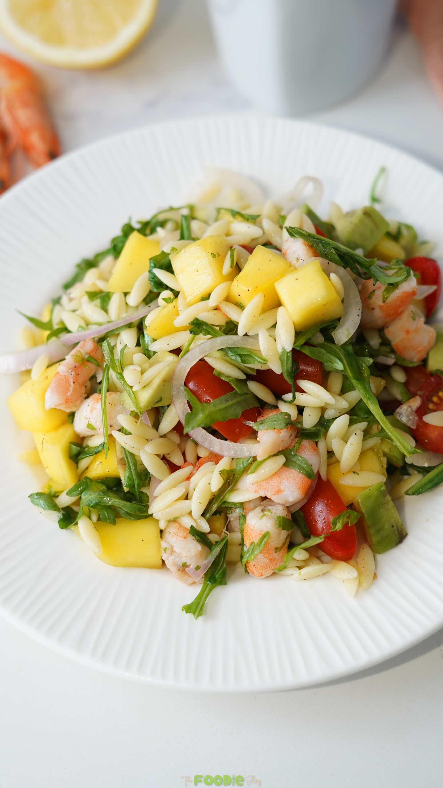 Prawn mango avocado summer salad served on a white plate with orzo, shrimp, mango, avocado, arugula, tomatoes, onion, and cilantro.