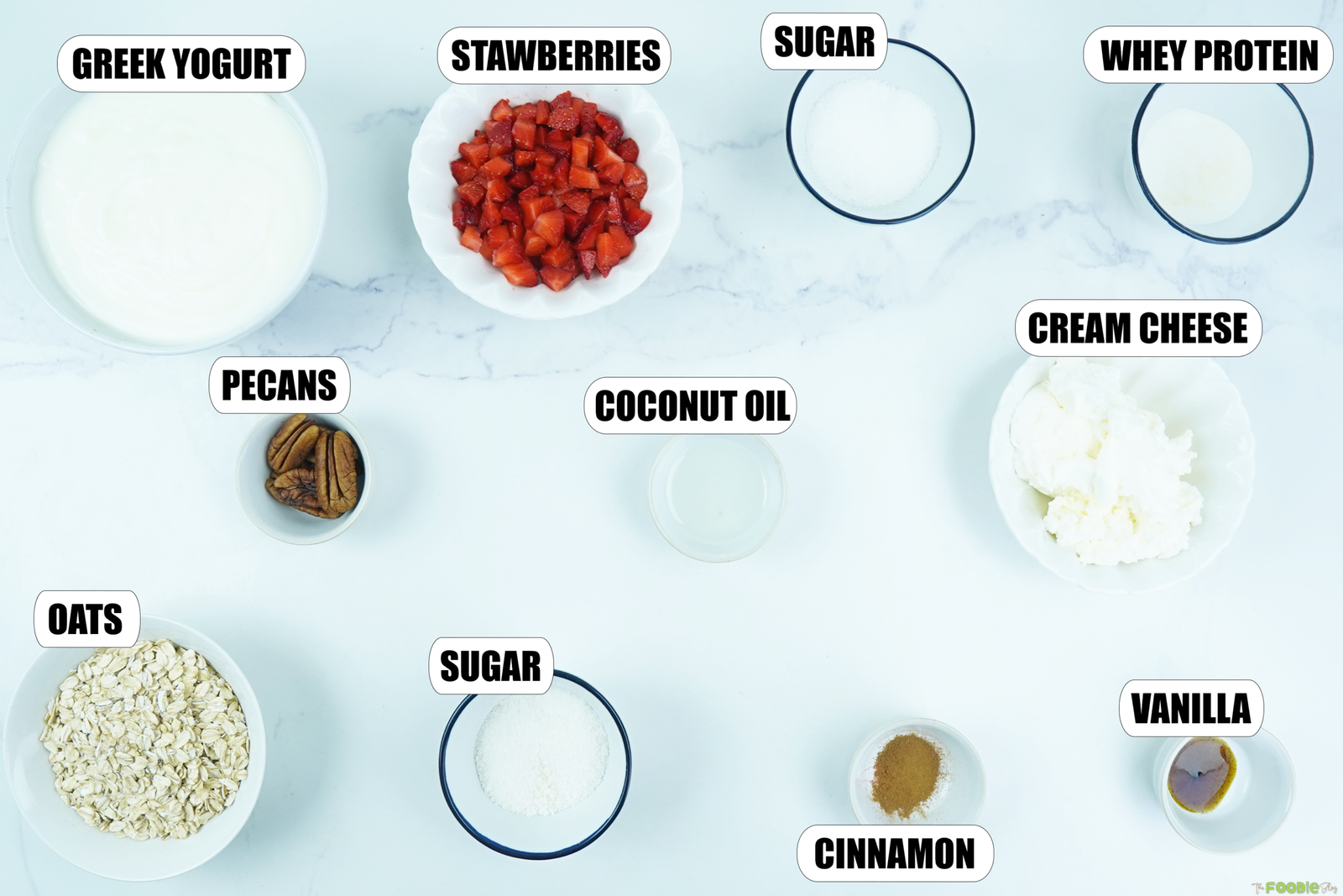 Ingredients for protein no bake cheesecake arranged on a white surface, including Greek yogurt, strawberries, oats, pecans, cream cheese, whey protein, vanilla, cinnamon, sweetener, and coconut oil.