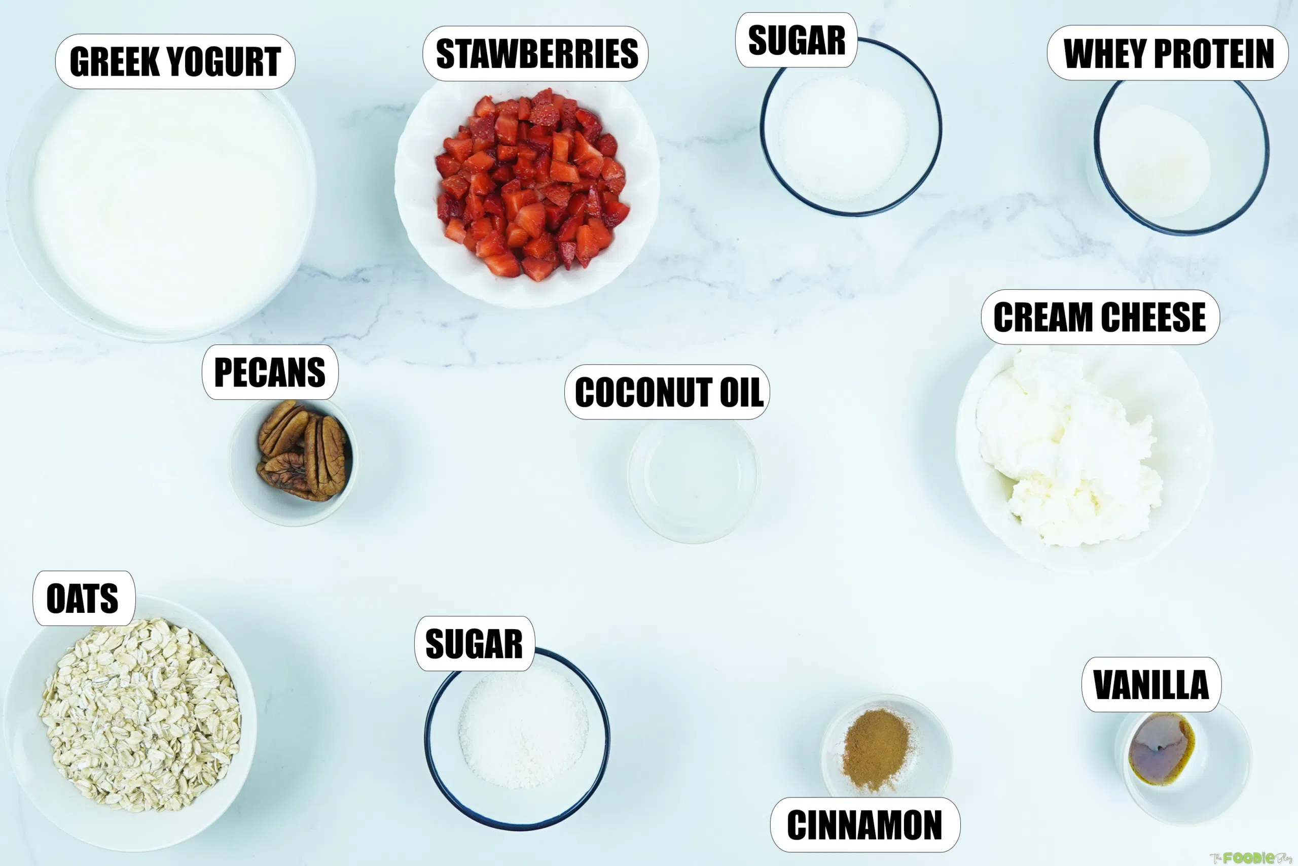 Ingredients for protein no bake cheesecake arranged on a white surface, including Greek yogurt, strawberries, oats, pecans, cream cheese, whey protein, vanilla, cinnamon, sweetener, and coconut oil.