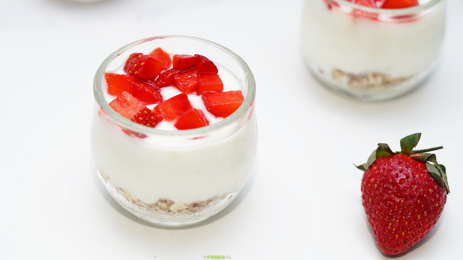 Small glass cups of creamy no bake protein cheesecake topped with chopped strawberries on a white surface.