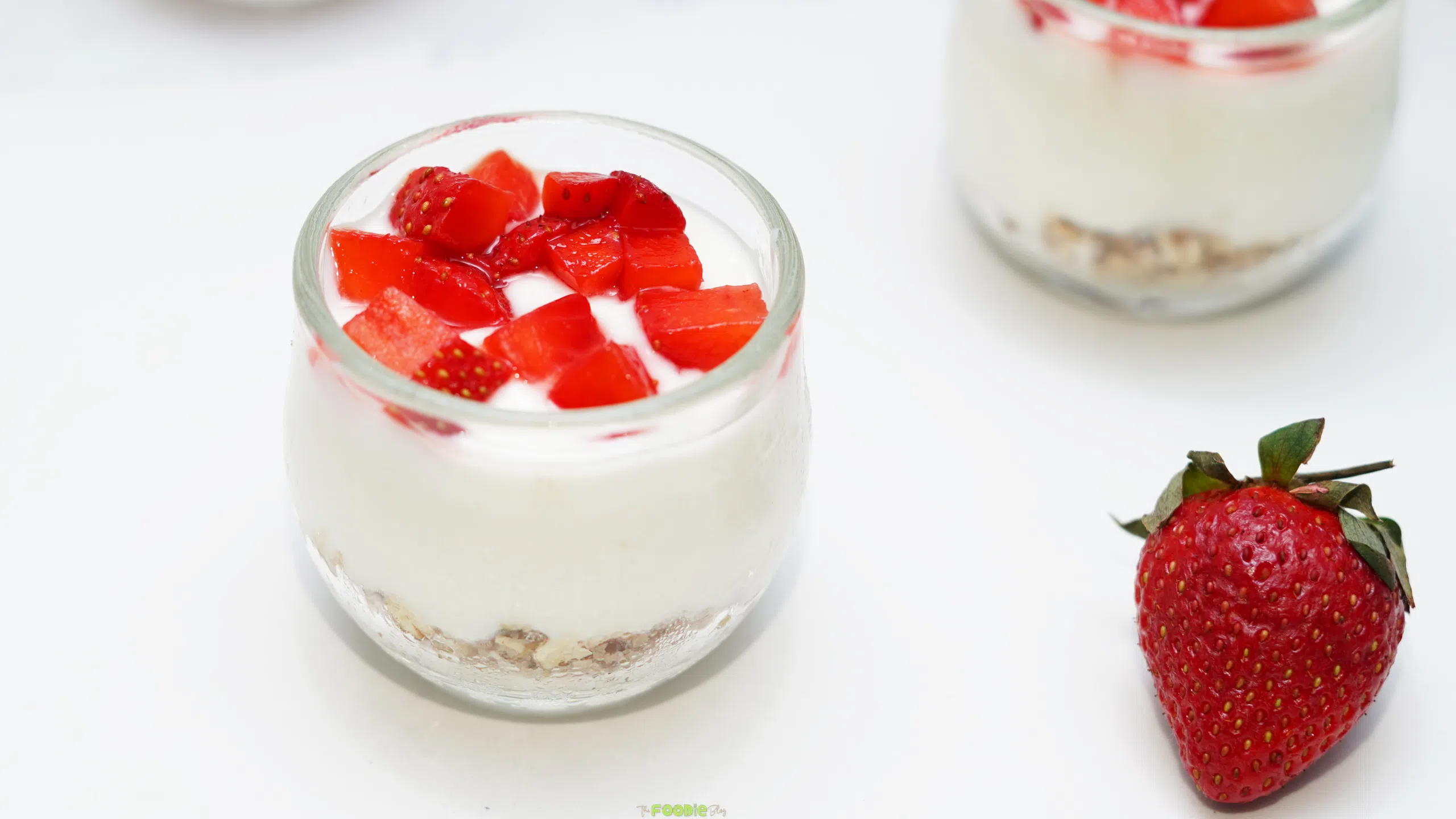 Small glass cups of creamy no bake protein cheesecake topped with chopped strawberries on a white surface.