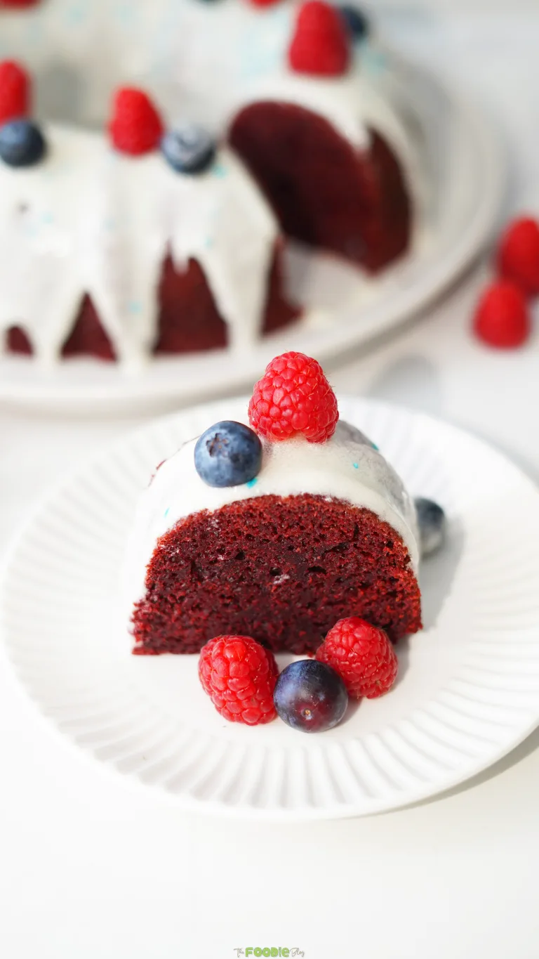 Slice of red velvet cake with cream cheese frosting, raspberries, and blueberries on a white plate.
