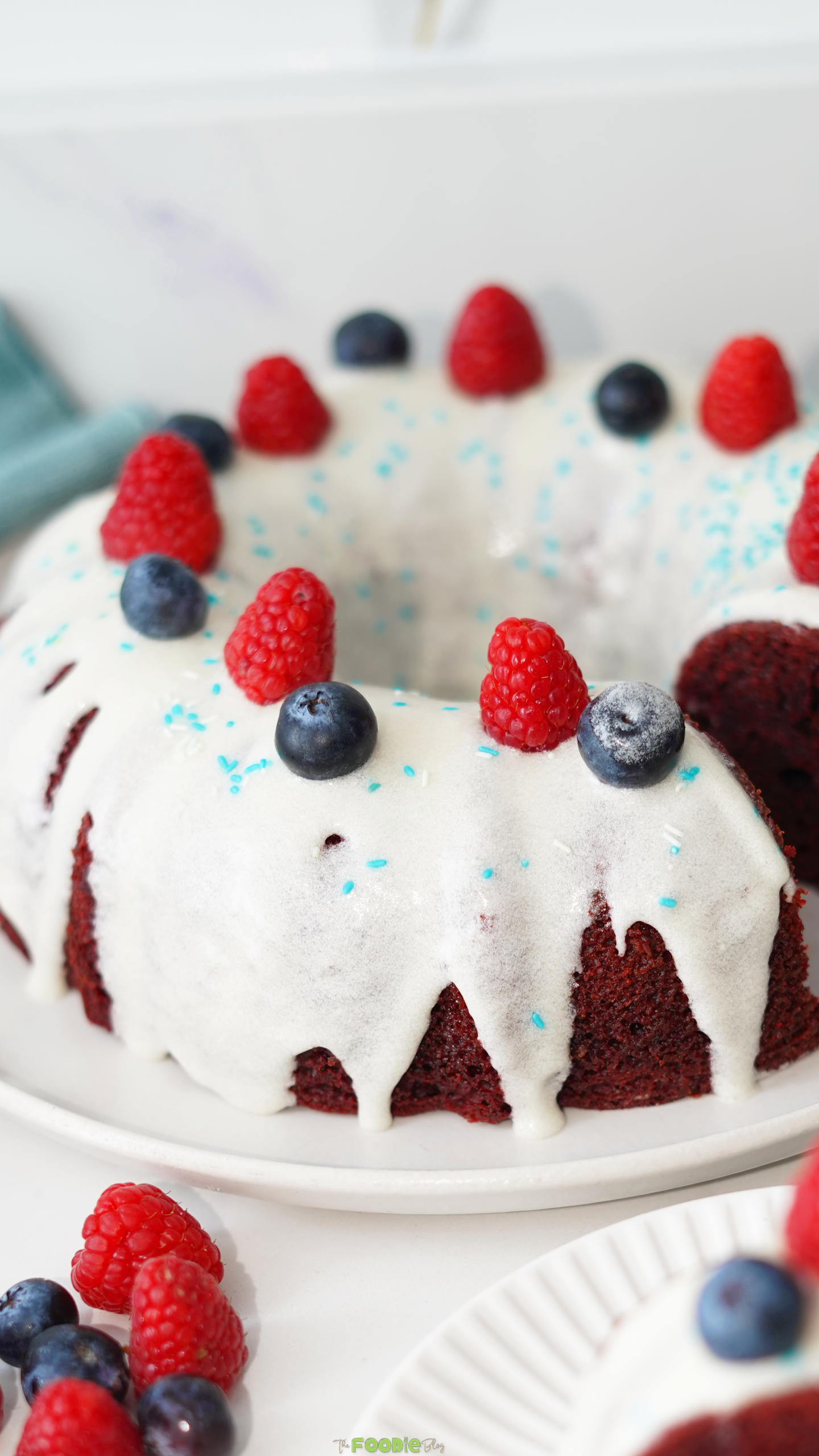 Patriotic red velvet Bundt cake decorated with raspberries, blueberries, cream cheese frosting, and sprinkles.