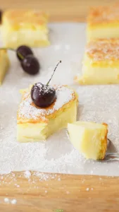 Vanilla custard cake square dusted with powdered sugar and topped with a fresh cherry.