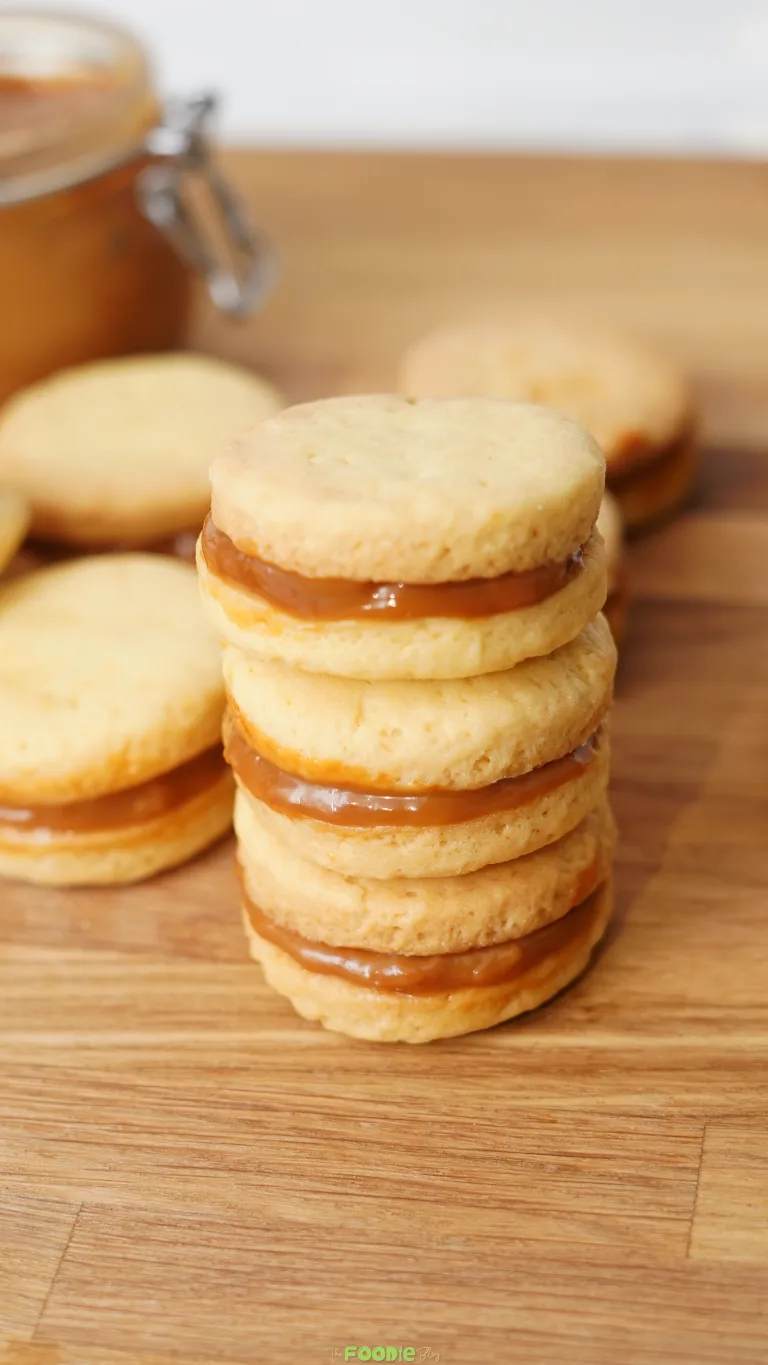 Stacked shortbread cookie sandwiches with dulce de leche filling on a wooden board.
