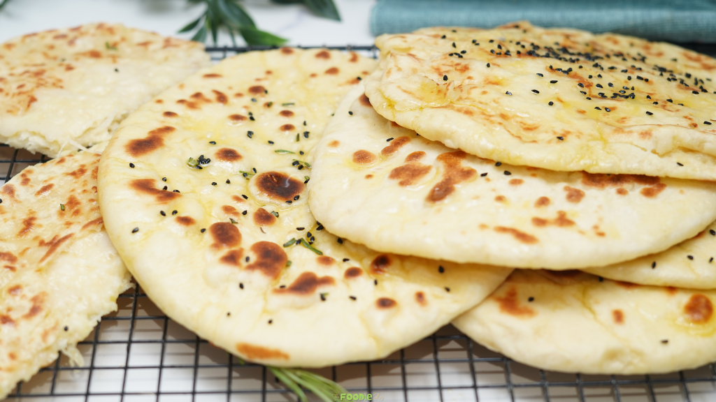 Homemade cheese naan with golden brown skillet spots, melted butter, and black sesame seeds arranged on a cooling rack.