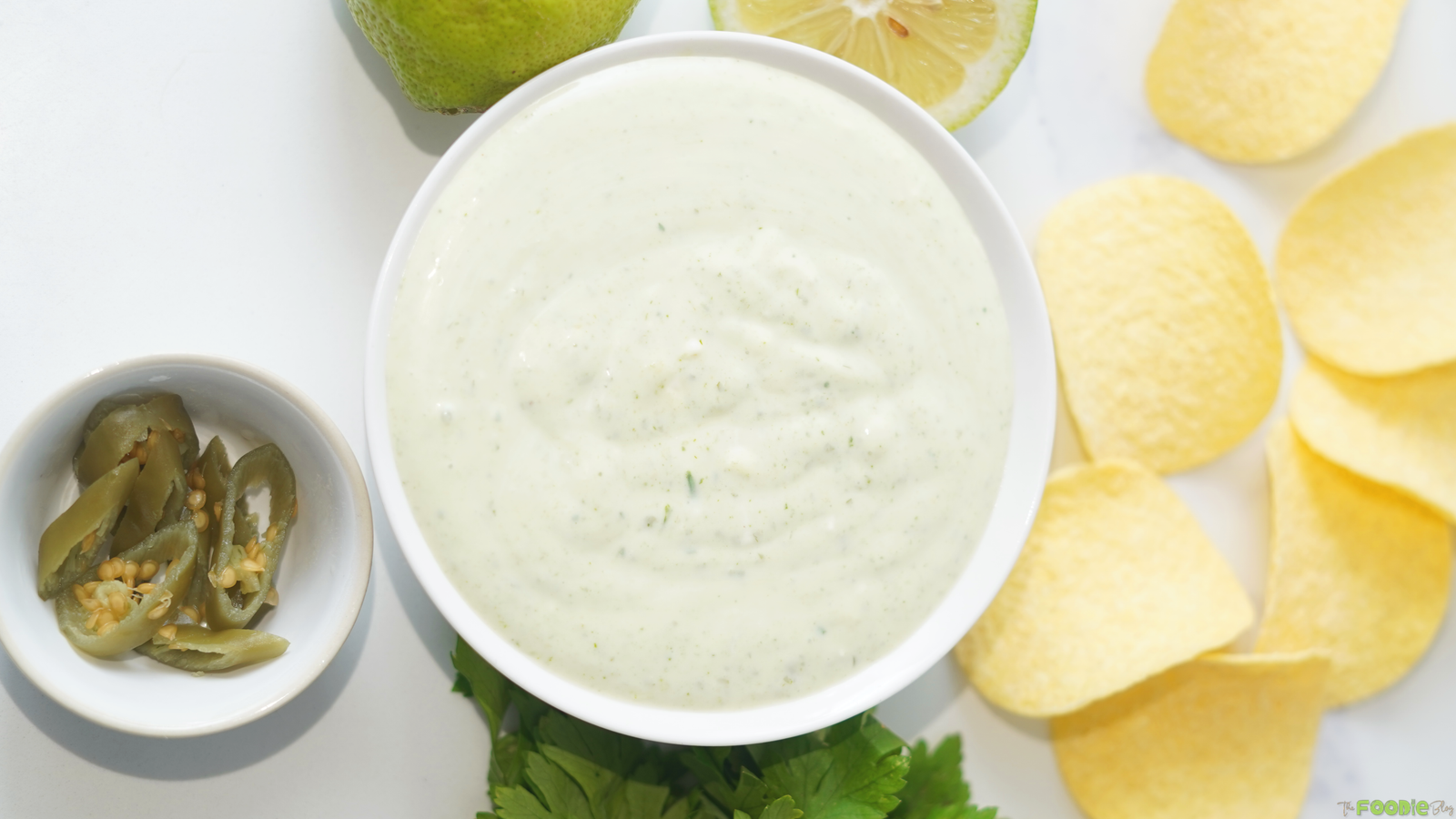 Overhead view of creamy jalapeño dip in a white bowl with potato chips, lime, cilantro, and pickled jalapeños.