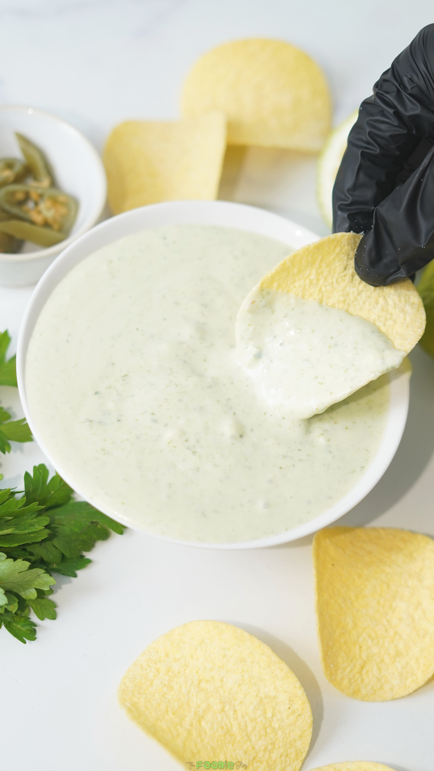 A potato chip dipped into creamy jalapeño ranch dip beside cilantro and pickled jalapeños.