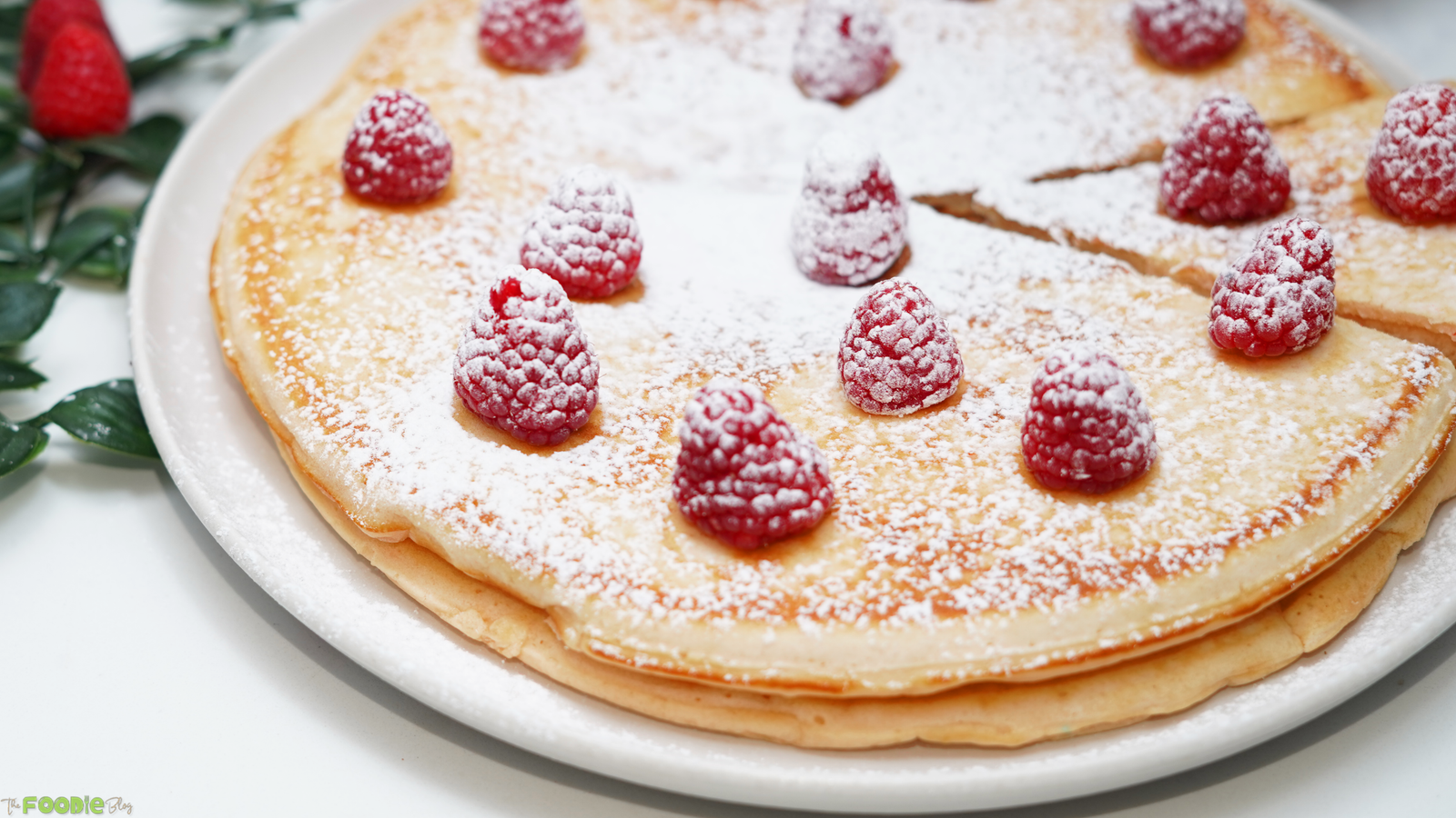 Close-up of golden Giant Pancakes stacked on a white plate with raspberries and powdered sugar.