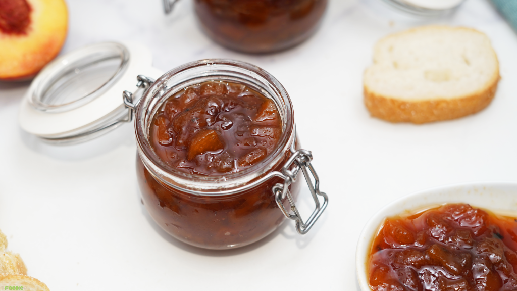 Jar of homemade peach preserves served with fresh peach and sliced bread.