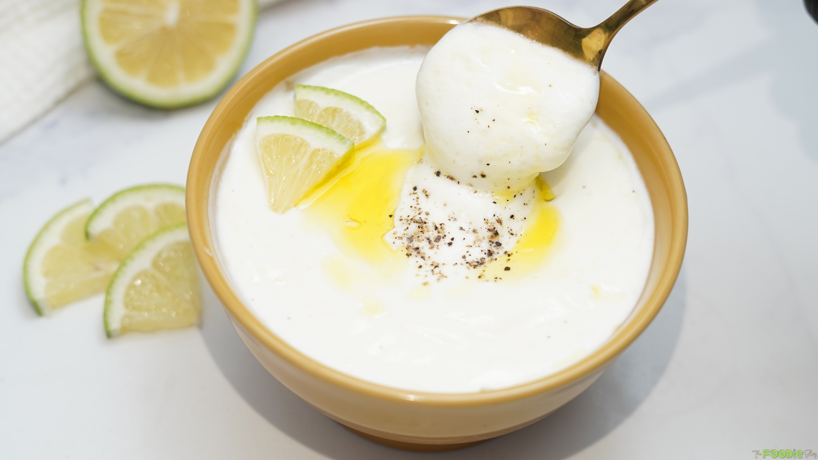 Spoon lifting creamy lemon yogurt sauce from a bowl with lemon wedges and olive oil.