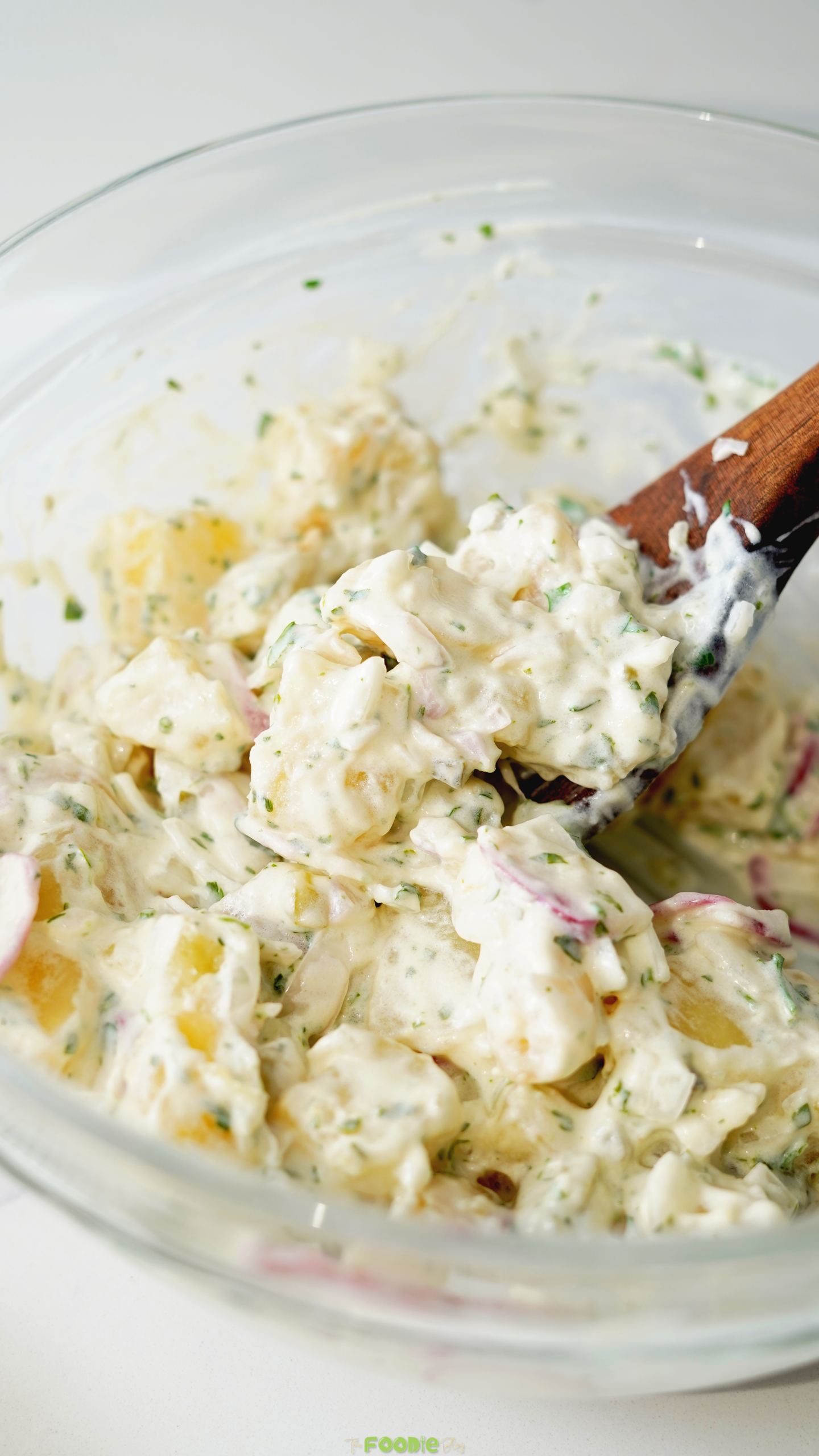 Potato Salad being mixed in a glass bowl with creamy dressing, potatoes, herbs, radishes, and onion.