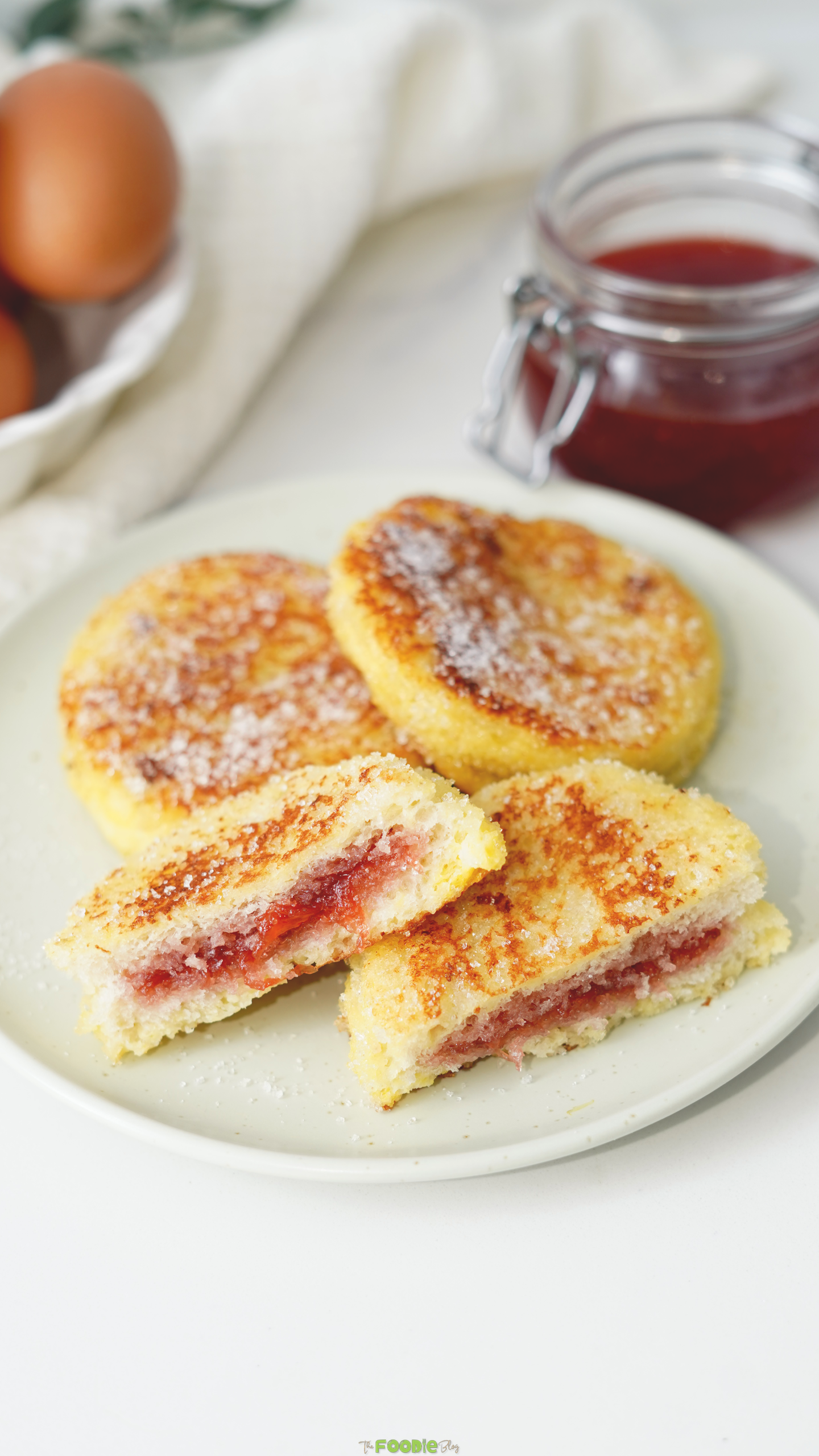 Sugar-coated French toast rounds with strawberry jam filling served beside eggs and a jar of jam.