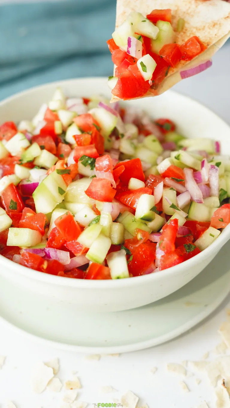 Tomato cucumber salad scooped onto a crispy pita chip above a bowl of fresh chopped salad.