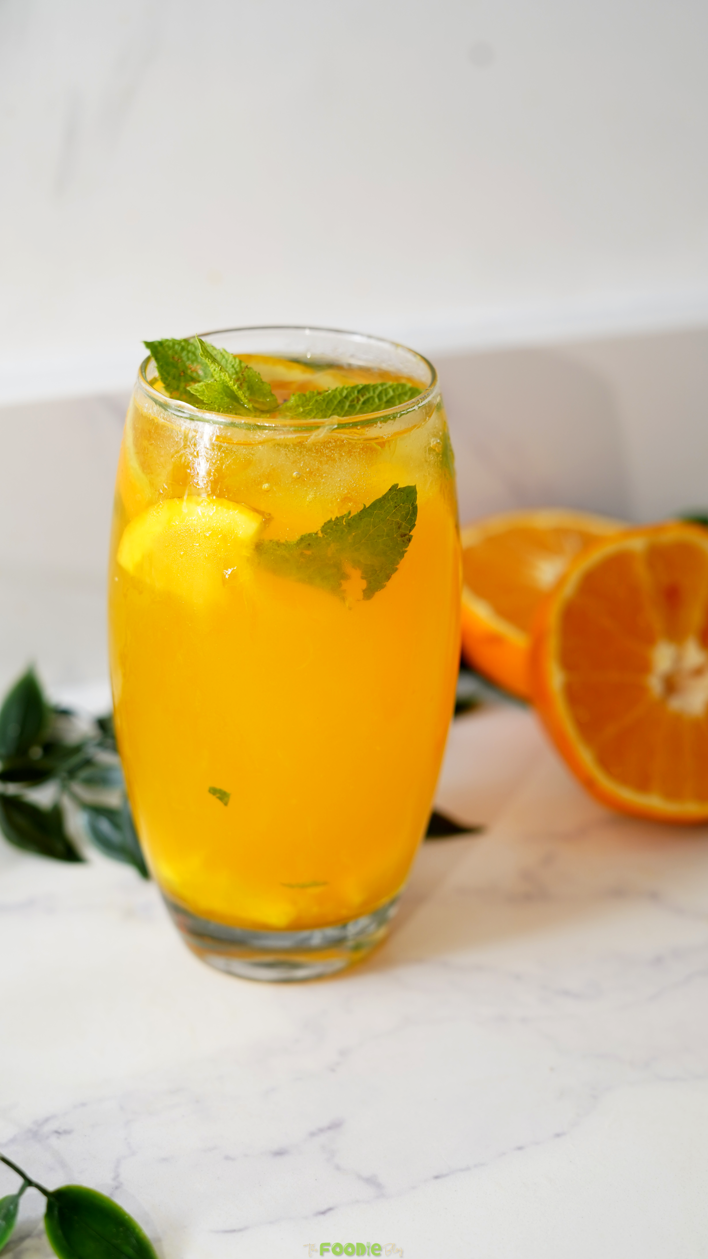 Tall glass of orange mojito with vodka, ice, fresh mint, and citrus slices beside fresh oranges.