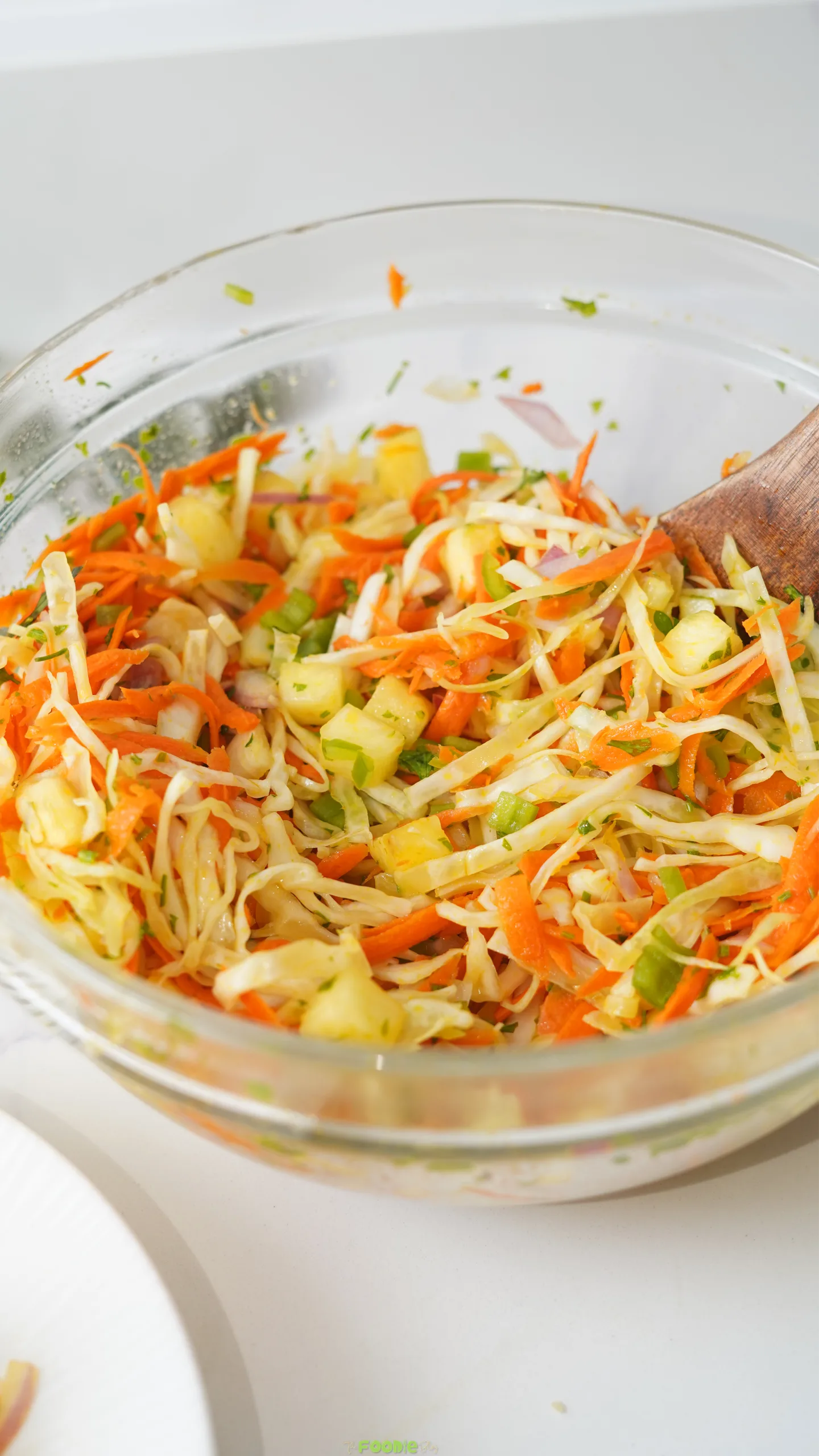 Pineapple cabbage slaw with shredded carrots, red onion, jalapeños, and cilantro being tossed in a glass bowl.