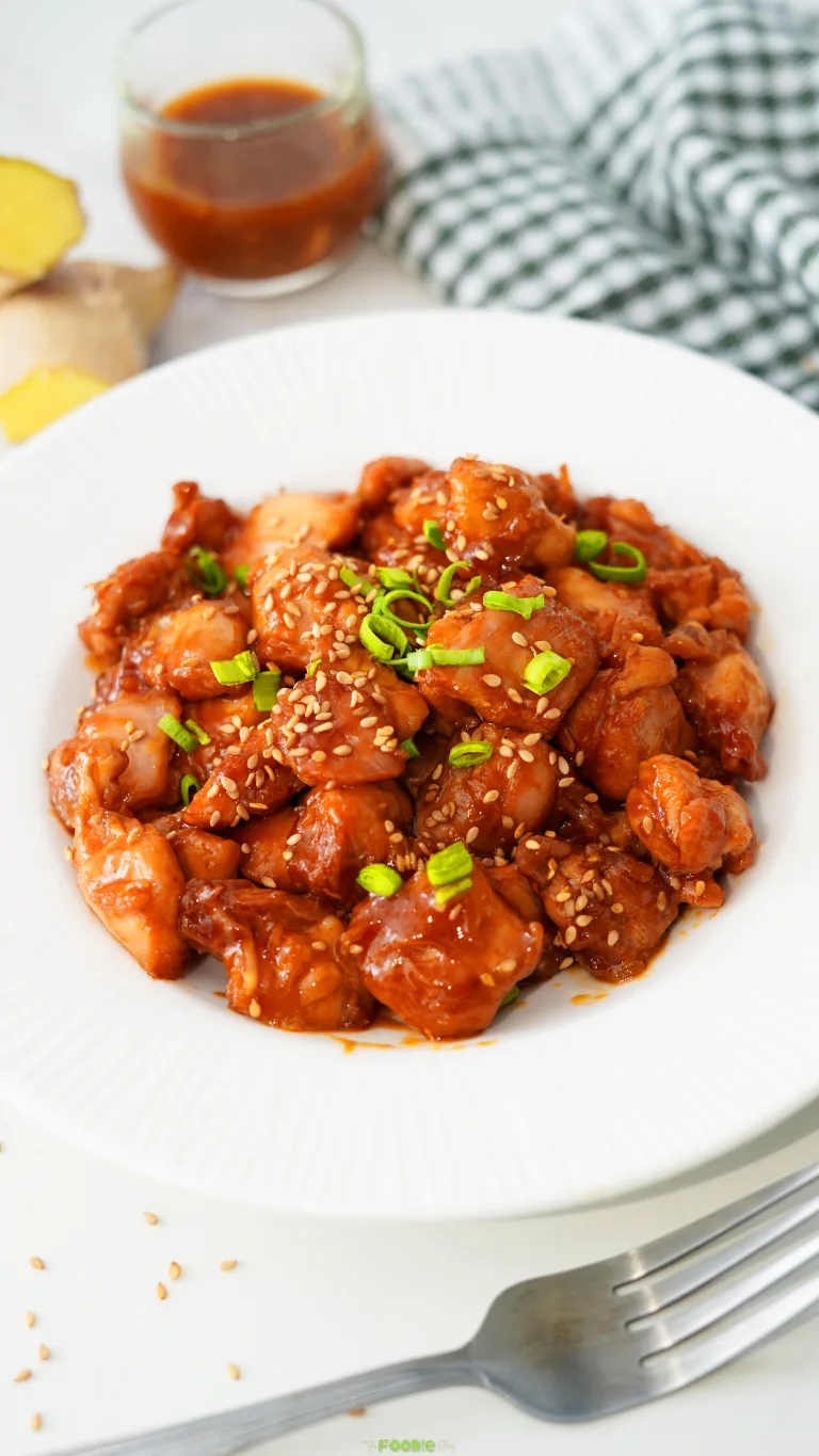 Sticky honey garlic chicken pieces served on a white plate with sesame seeds and sliced green onions.