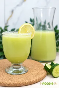 Fresh cucumber lemonade served in a glass with a lemon slice, with a pitcher and sliced cucumber in the background.