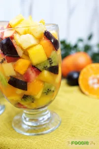 Fresh fruit salad served in glass cups with pineapple, mango, kiwi, plums, apples, persimmons, and orange.
