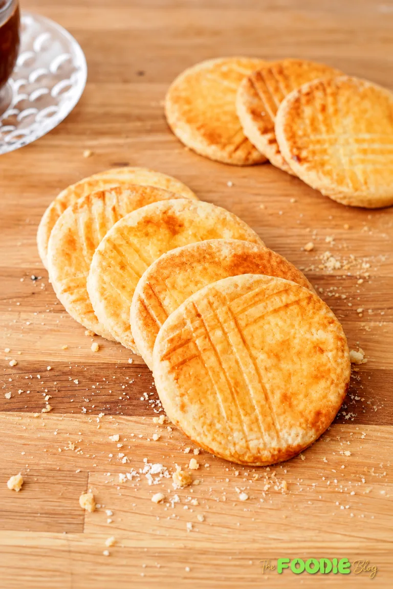 Stack of golden Galettes Bretonnes with several cookies leaning beside it and crumbs scattered on a wooden board.