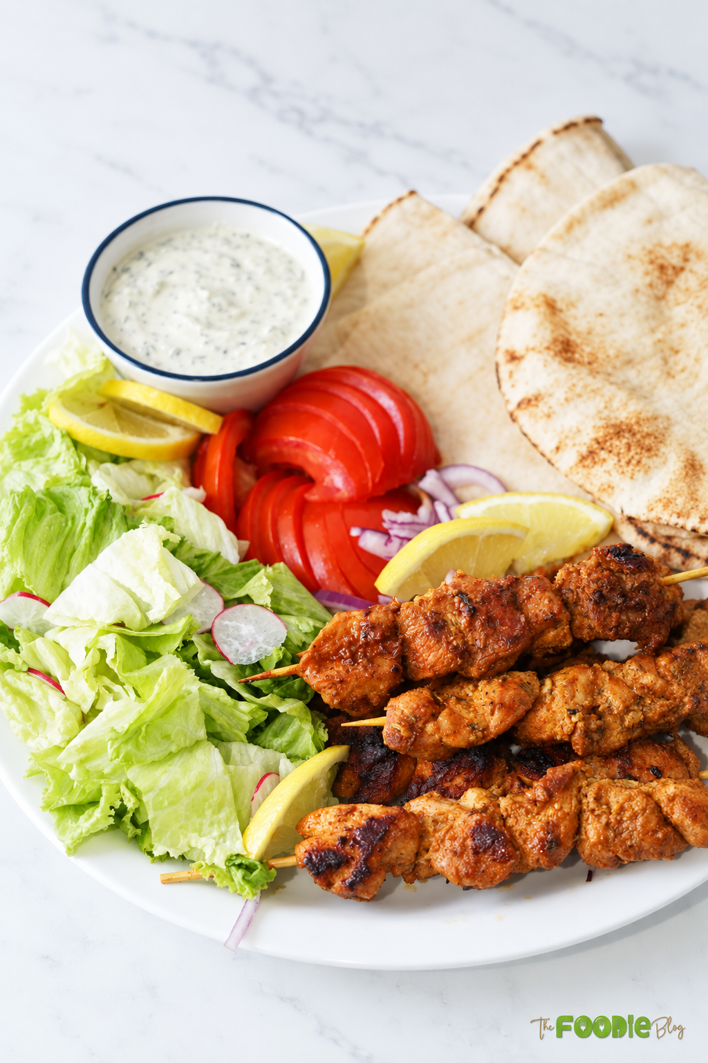 Cooked chicken skewers served with lettuce, tomatoes, pita bread, lemon wedges, and creamy herb sauce.