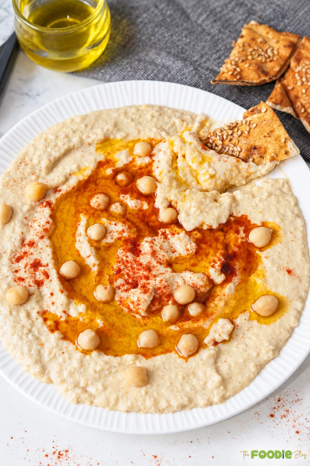 Creamy hummus spread on a plate with olive oil, whole chickpeas, paprika, and pita chips