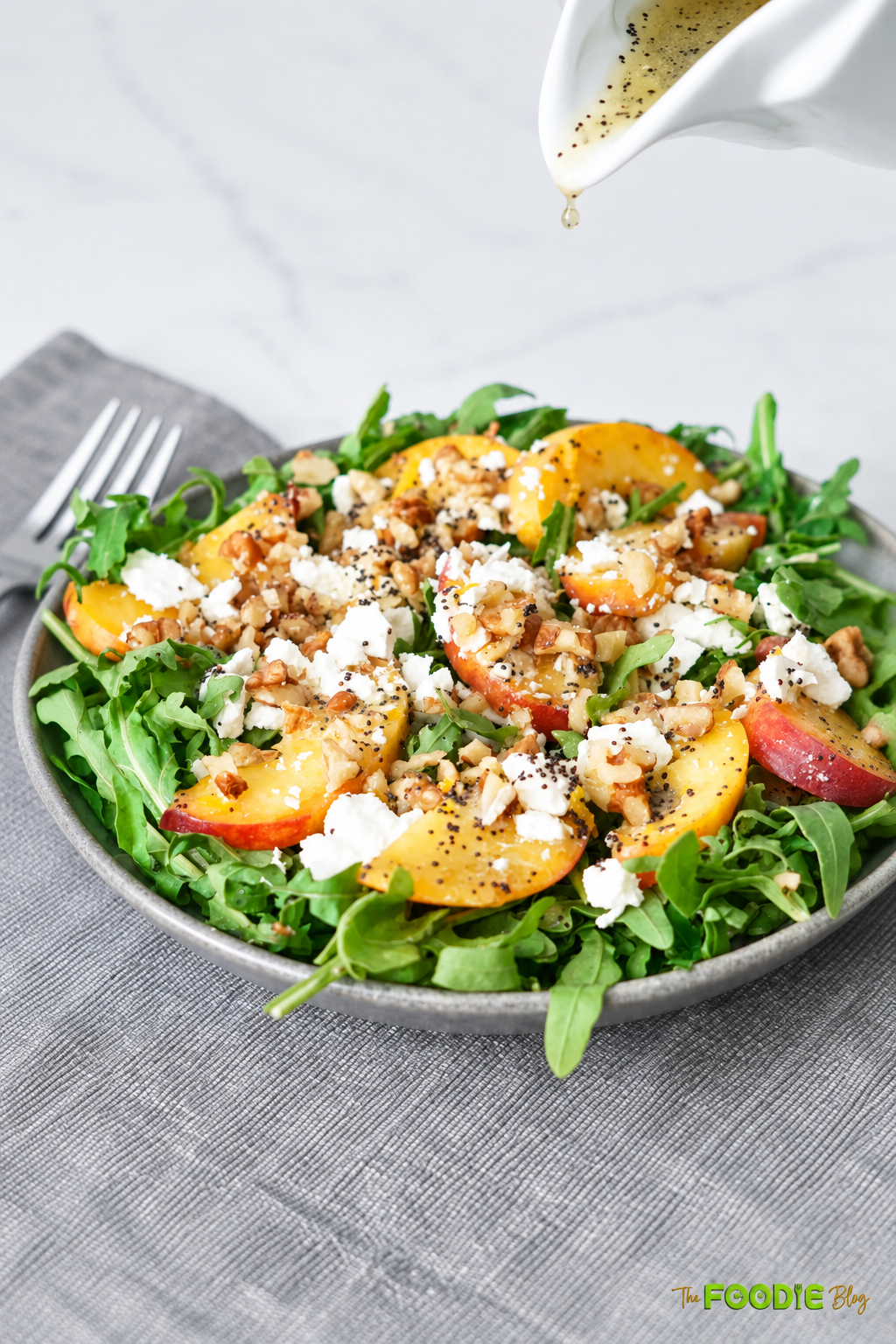 Homemade poppy seed dressing being drizzled over peach arugula salad with feta and walnuts.