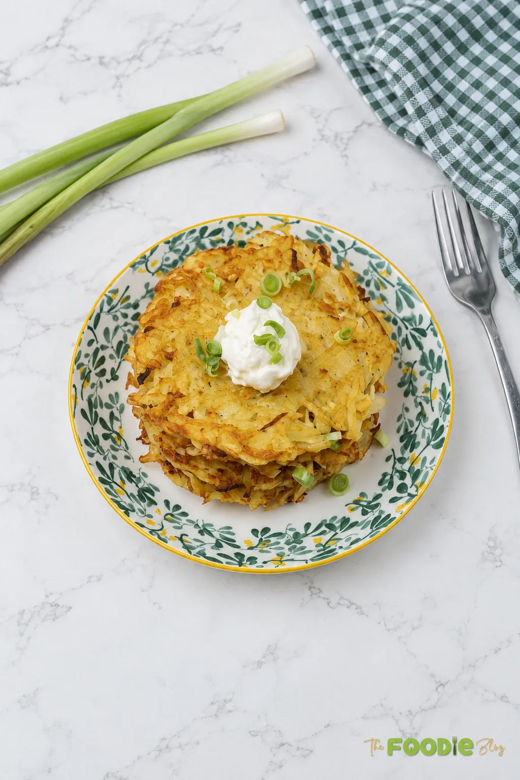 German potato pancakes stacked on a patterned plate with sour cream and sliced green onions.