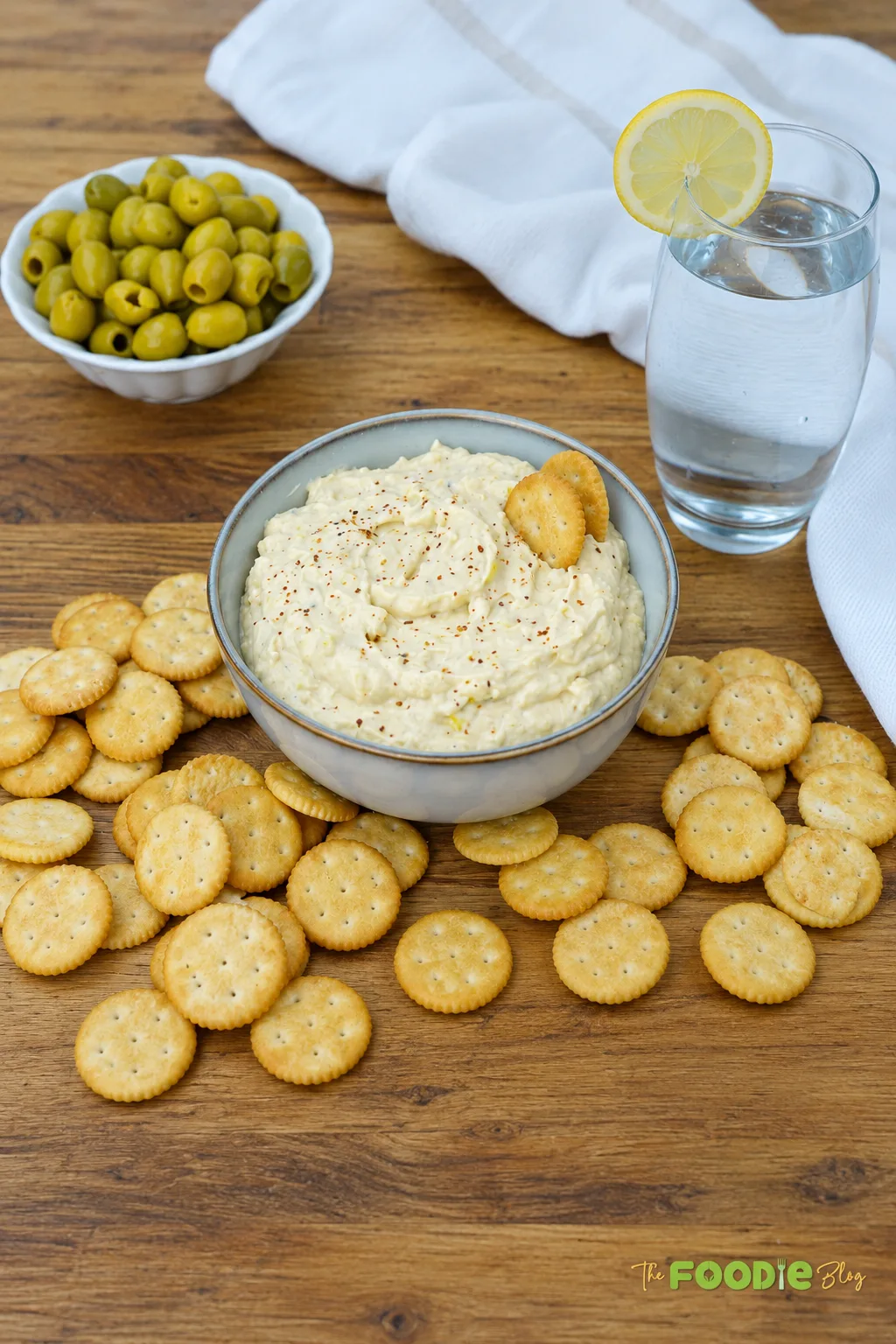 Bowl of creamy green olive dip surrounded by crackers with green olives on a wooden table.